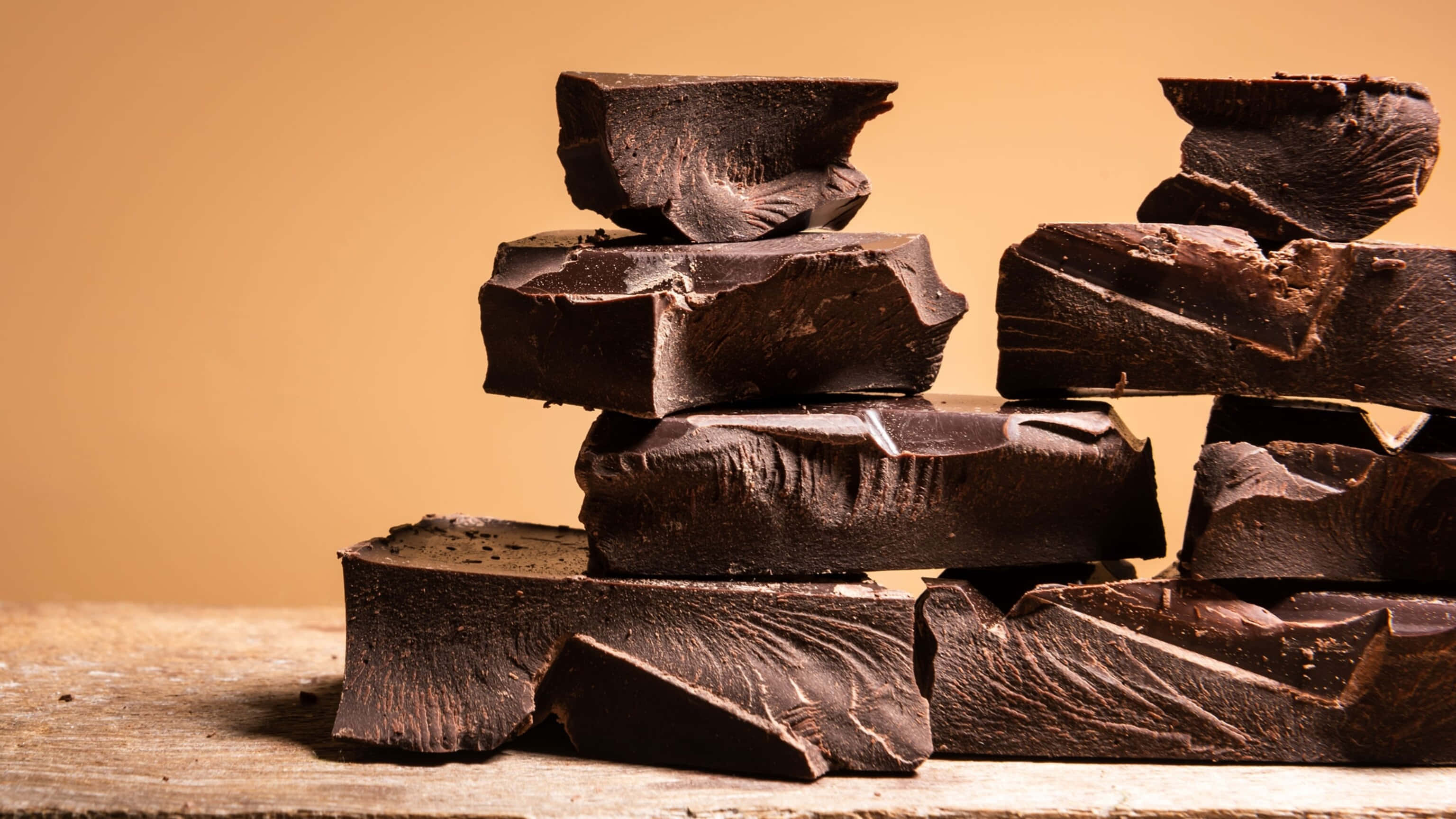 Dark Chocolate 桌布 壁紙 — 濃郁的黑巧克力塊不均勻地堆疊在質樸的木質表面上，背景是溫暖的米色。