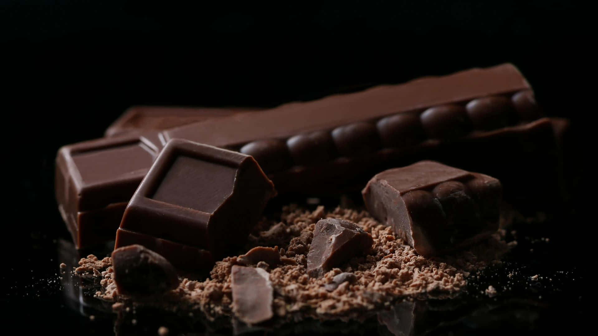 Dark Chocolate 桌布 壁紙 — 濃郁的黑巧克力條和碎塊，可可粉散落在光亮的黑色表面上，在戲劇性的燈光下。