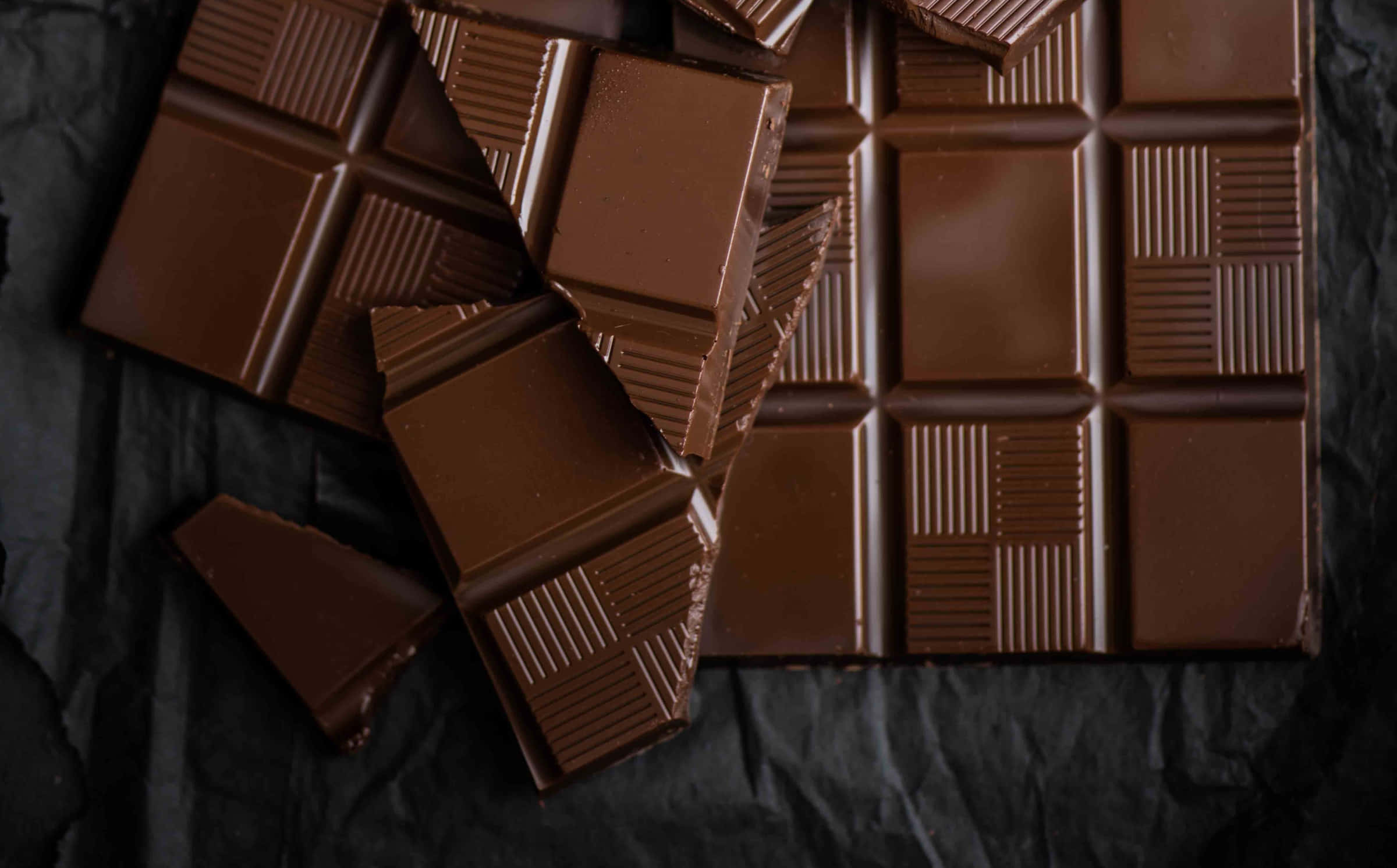Brown Chocolate 桌布 壁紙 — 濃郁的黑巧克力條和碎塊放在皺巴巴的黑色紙上，帶有光澤質感和深棕色調。