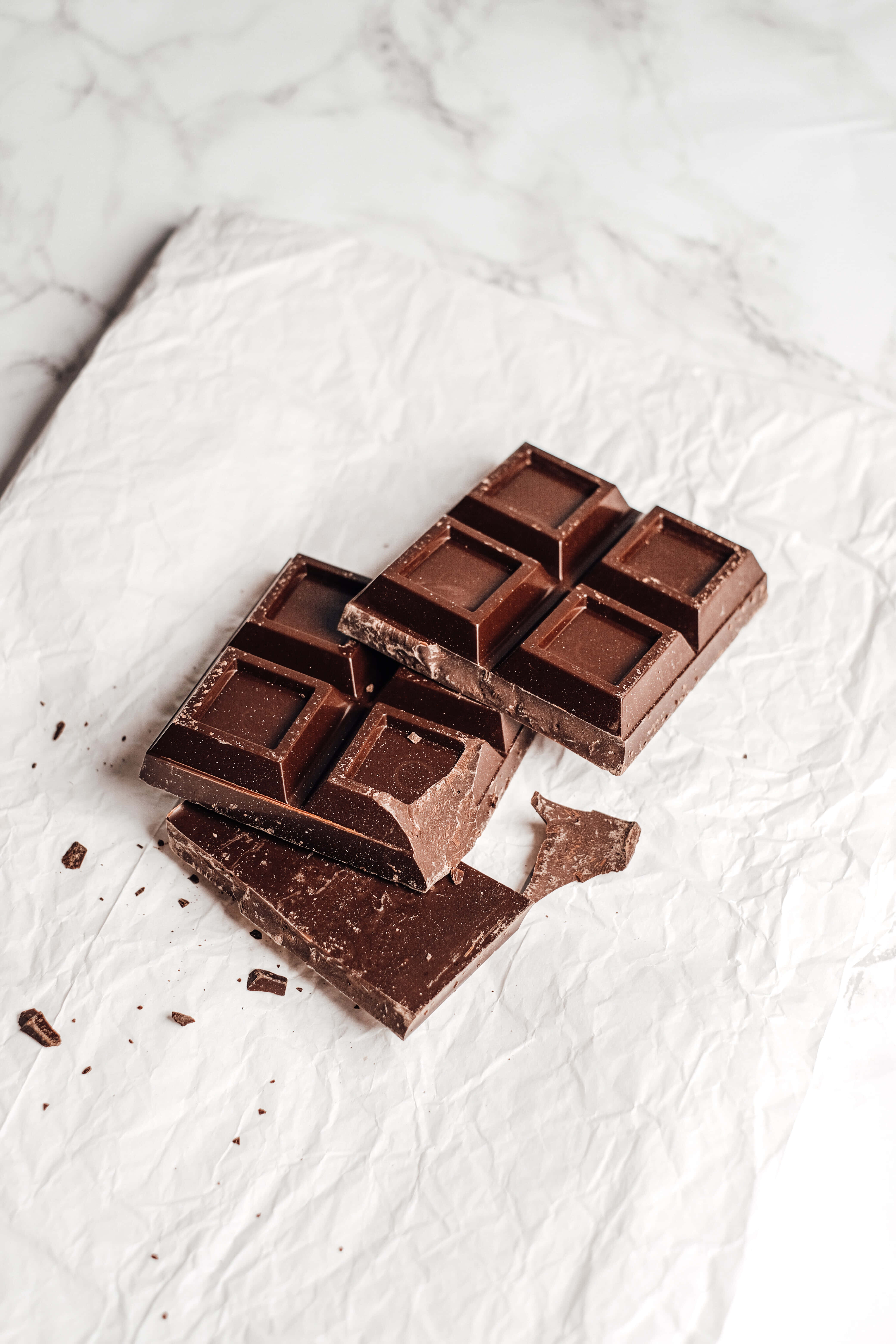 Dark Chocolate 桌布 壁紙 — 濃郁的黑巧克力棒碎成方塊，放在皺巴巴的白紙上，以大理石為背景。