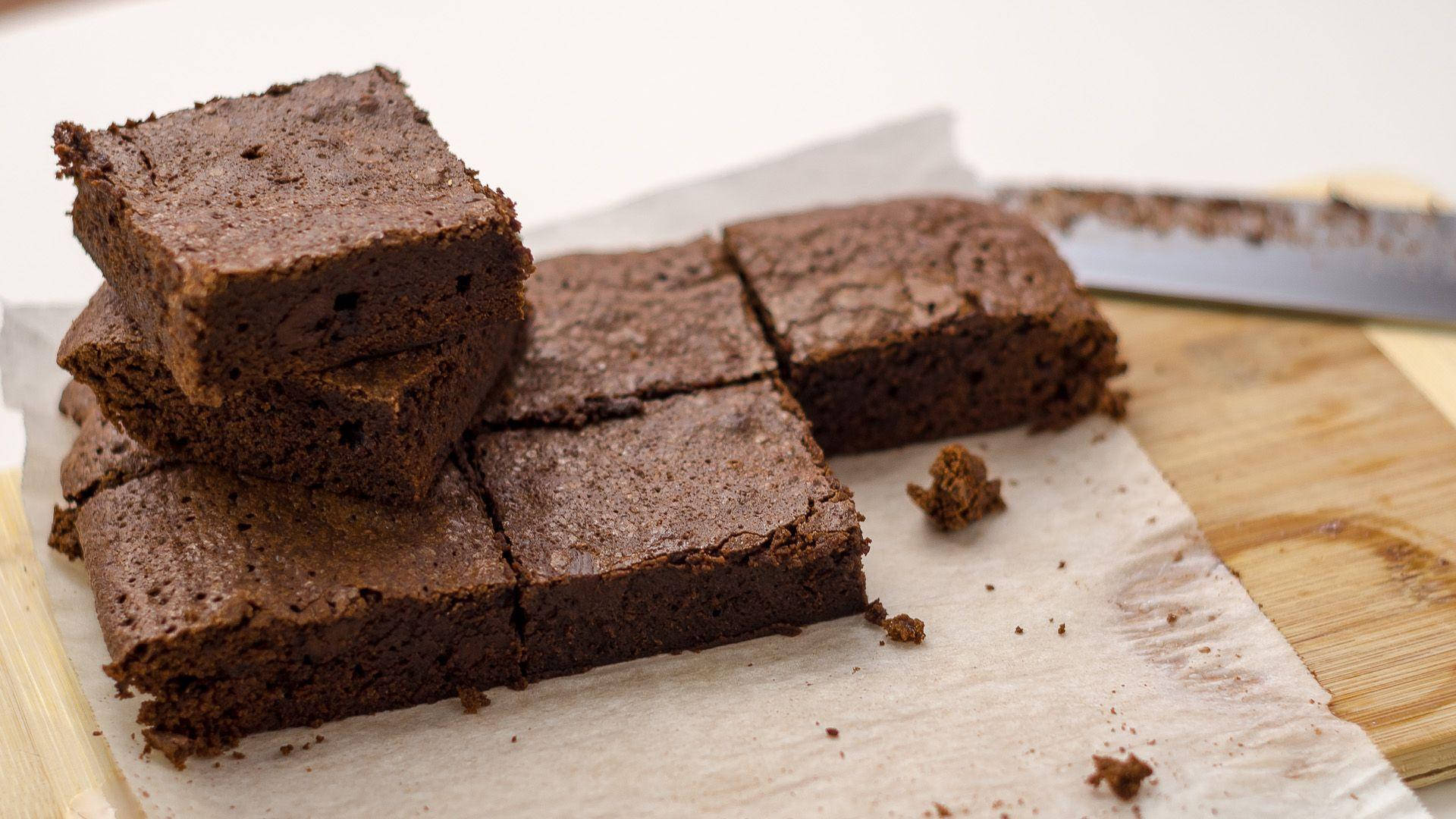 Brownies 桌布 壁紙 — 濃郁軟心巧克力布朗尼切成方塊，放在木砧板上的烘焙紙上，旁邊有刀。