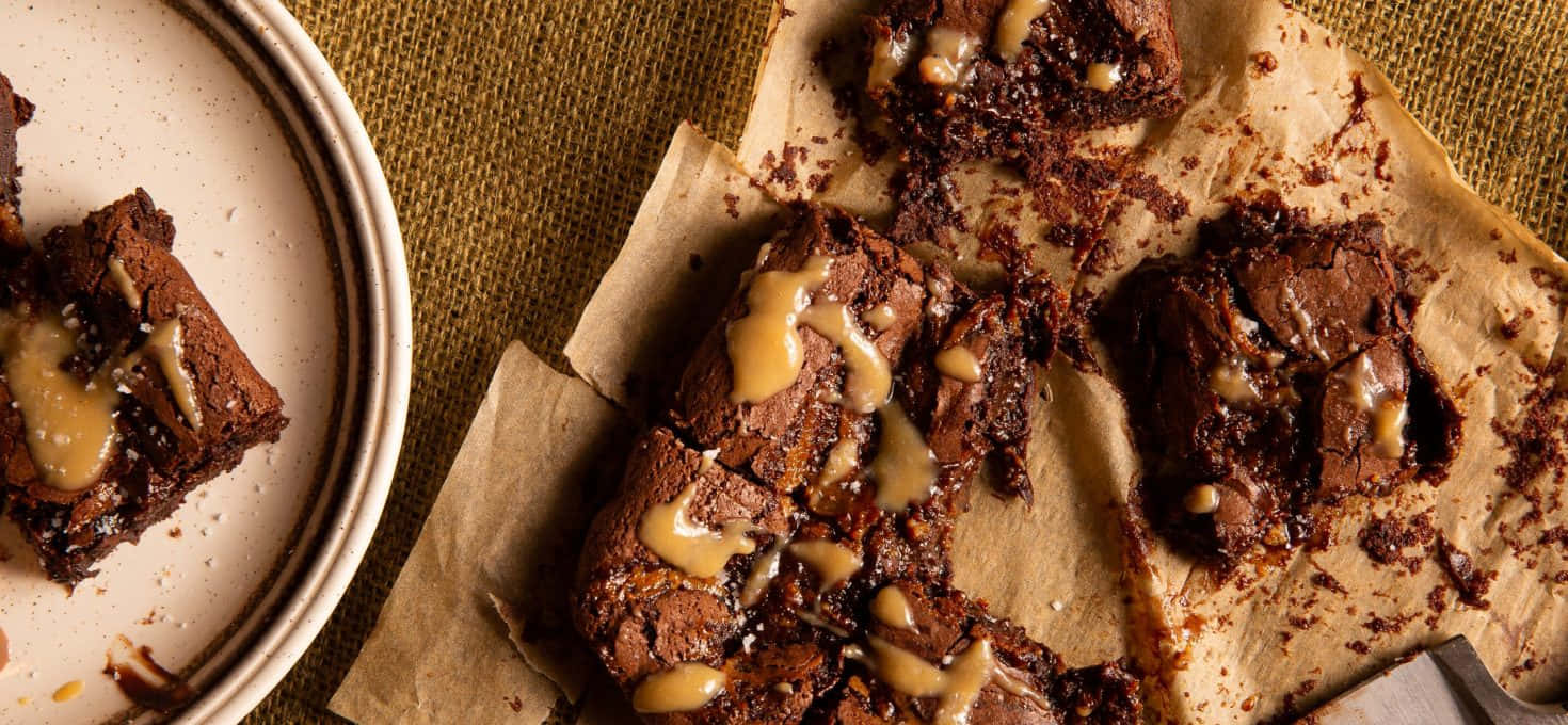 Caramel Brownie 桌布 壁紙 — 濃郁軟心巧克力布朗尼淋上焦糖醬，放在烘焙紙上，旁邊有一個盤子。