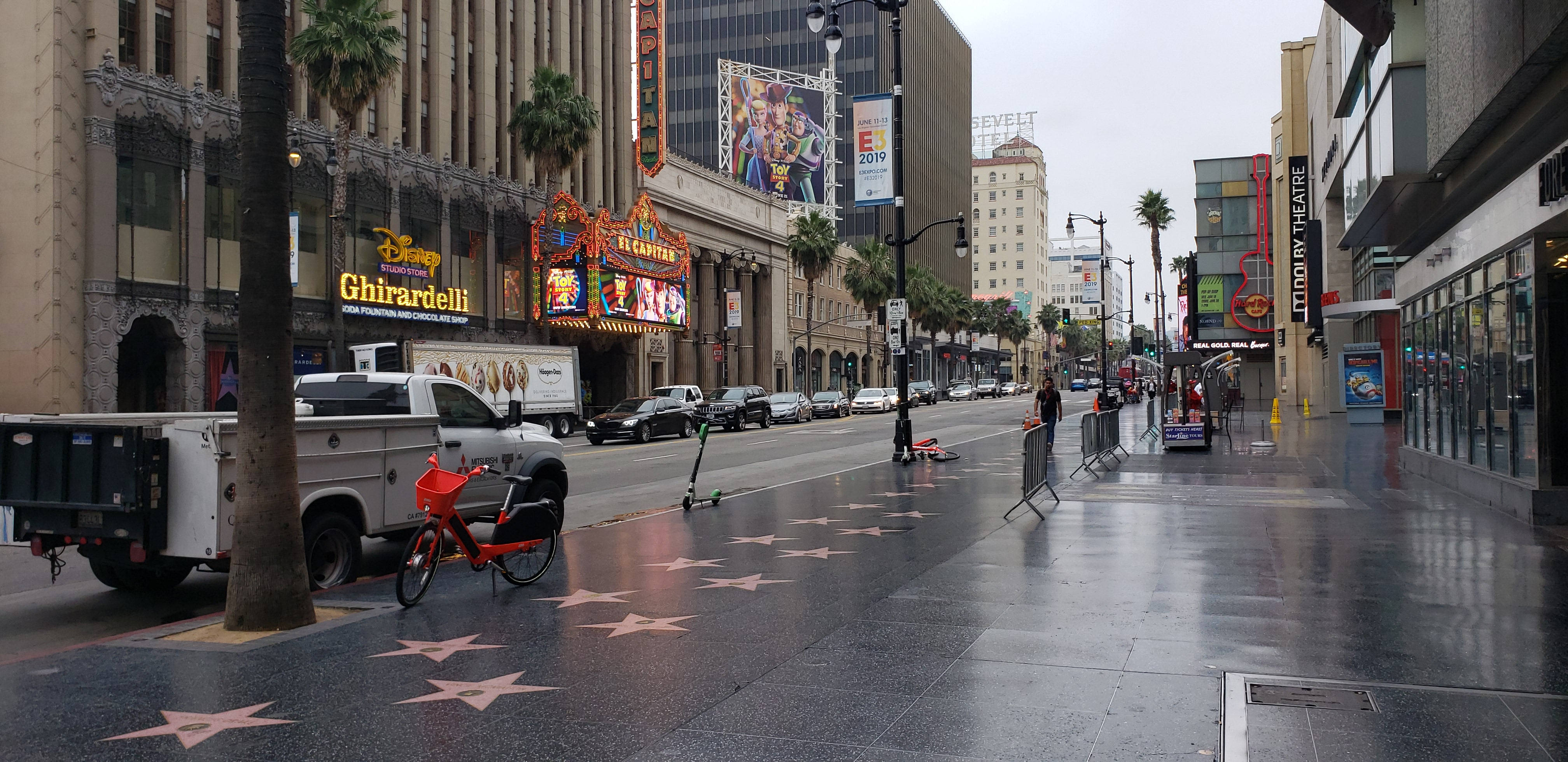 walk 桌布 壁紙 — 濕漉漉的好萊塢星光大道星星在陰天天空下閃爍，棕櫚樹排列在繁忙的街道上，兩旁是標誌性劇院和停泊的車輛。