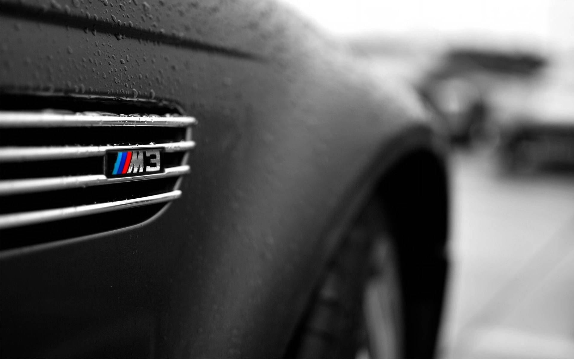 m 桌布 壁紙 — 濕潤的BMW M3徽章特寫，在柔和光線下閃爍，雨滴依附在汽車光滑的黑色表面。