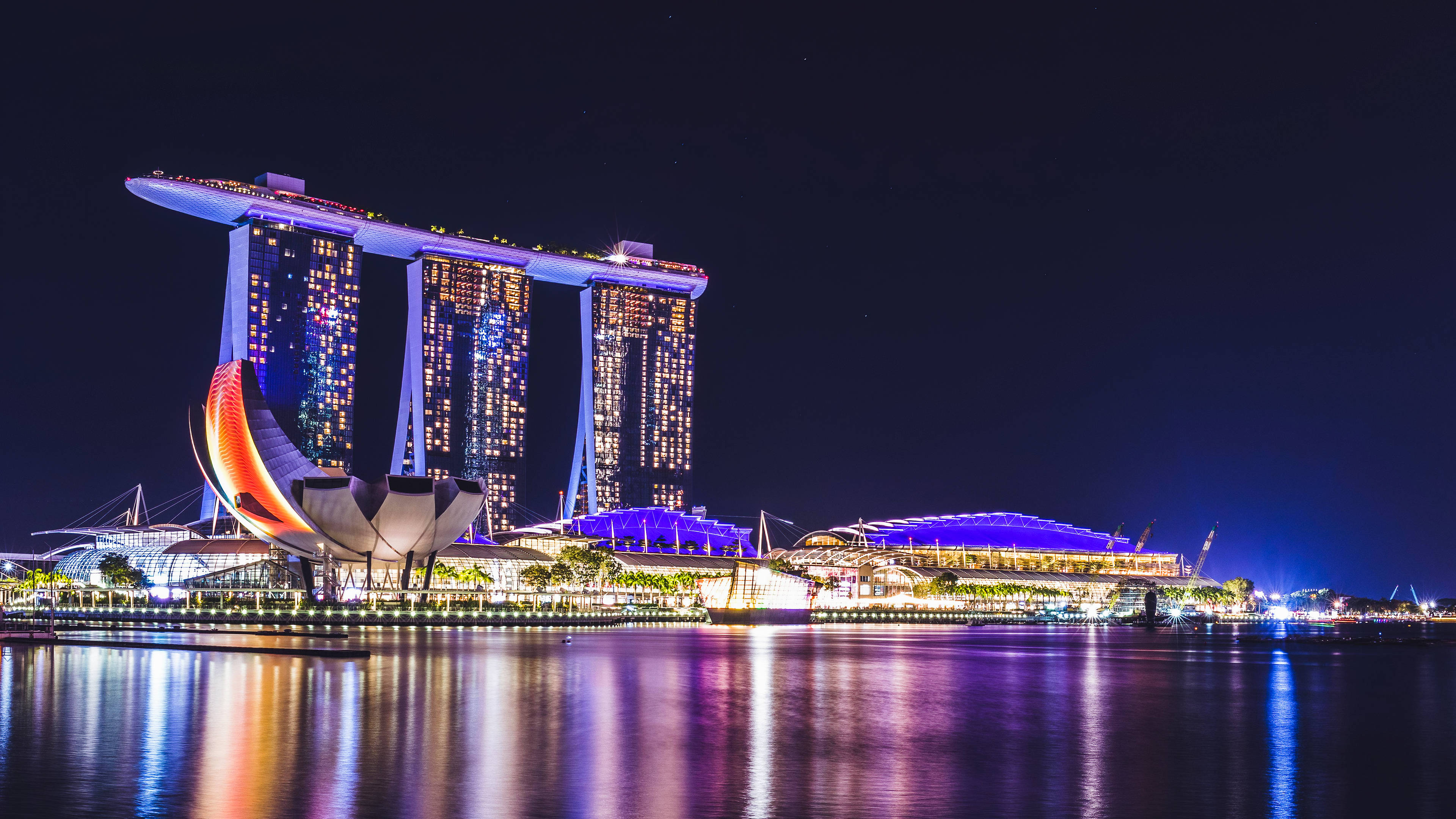 風景動畫 桌布 壁紙 — 濱海灣金沙在夜晚閃爍的海濱上空聳立，鮮豔的燈光倒映在平靜的水面上。