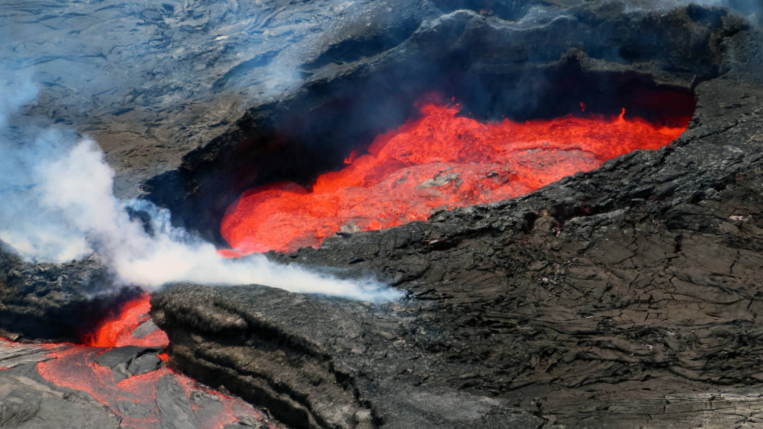 Volcano 桌布 壁紙 — 火山口噴發，熾熱的紅色熔岩流過黑暗崎嶇的地形，並冒出滾滾白煙。