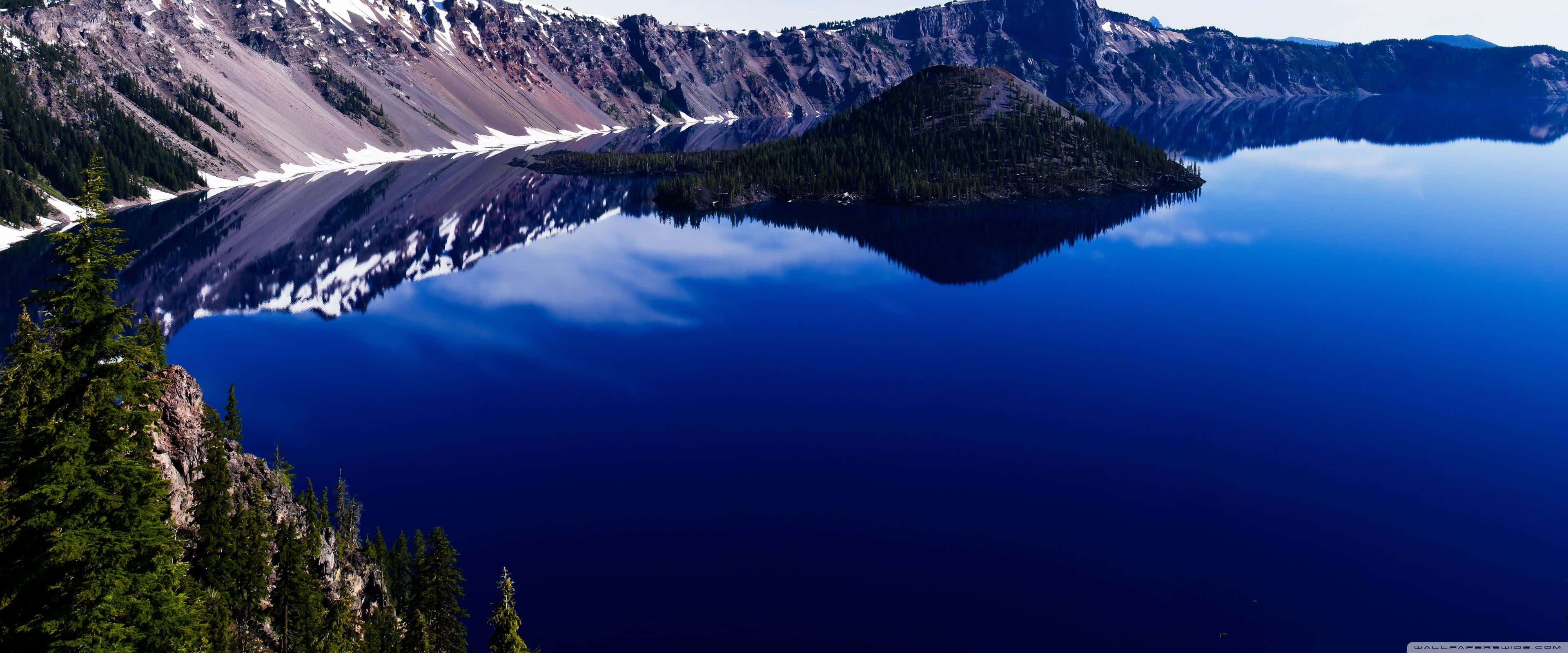 Apple 3840x1600 桌布 壁紙 — 火山口湖的壯麗全景，深藍色的湖水完美地映照出白雪覆蓋的山脈和常綠森林。