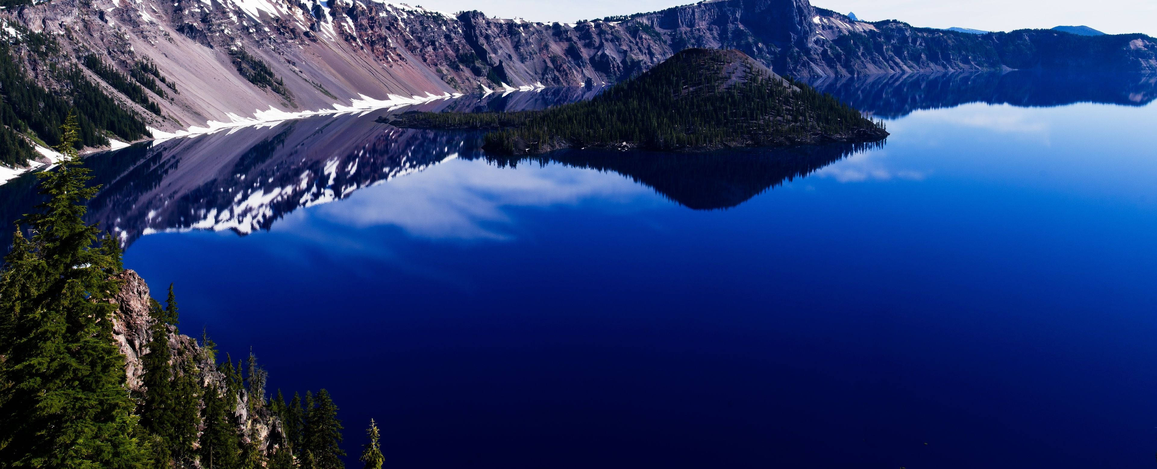 3840x1600 桌布 壁紙 — 火山口湖的壯麗景色，深藍色的湖水映照著白雪覆蓋的山峰和常綠森林，天空晴朗。