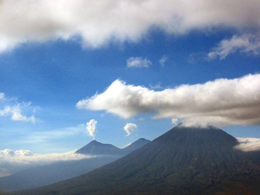 Guatemala 桌布 壁紙 — 火山在蔚藍天空下穿過雲層雄偉地升起，縷縷白雲在上空飄蕩。