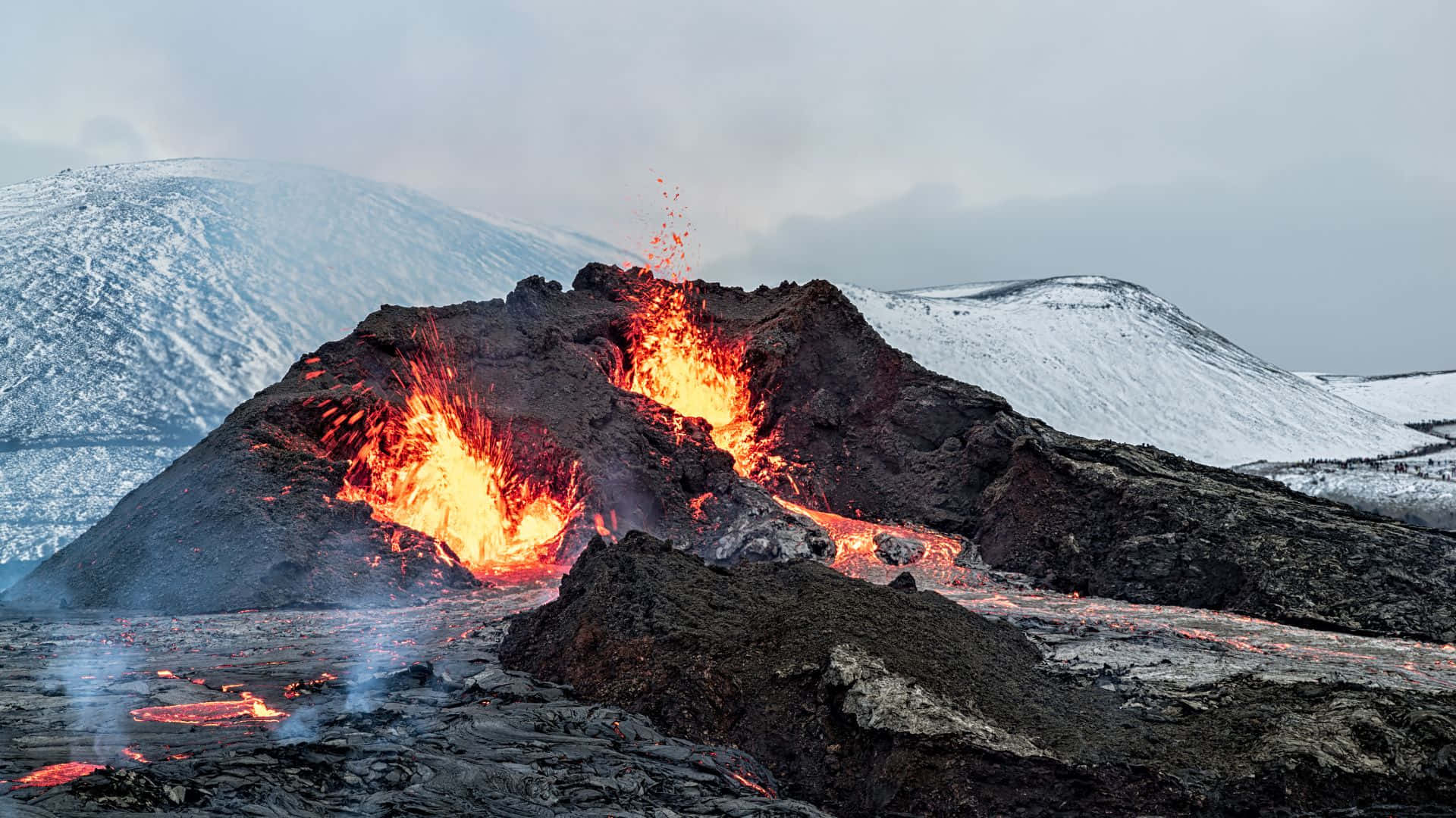 冰島 桌布 壁紙 — 火山爆發噴出熾熱的熔岩和灰燼，背景是白雪覆蓋的山脈，天空灰濛濛。