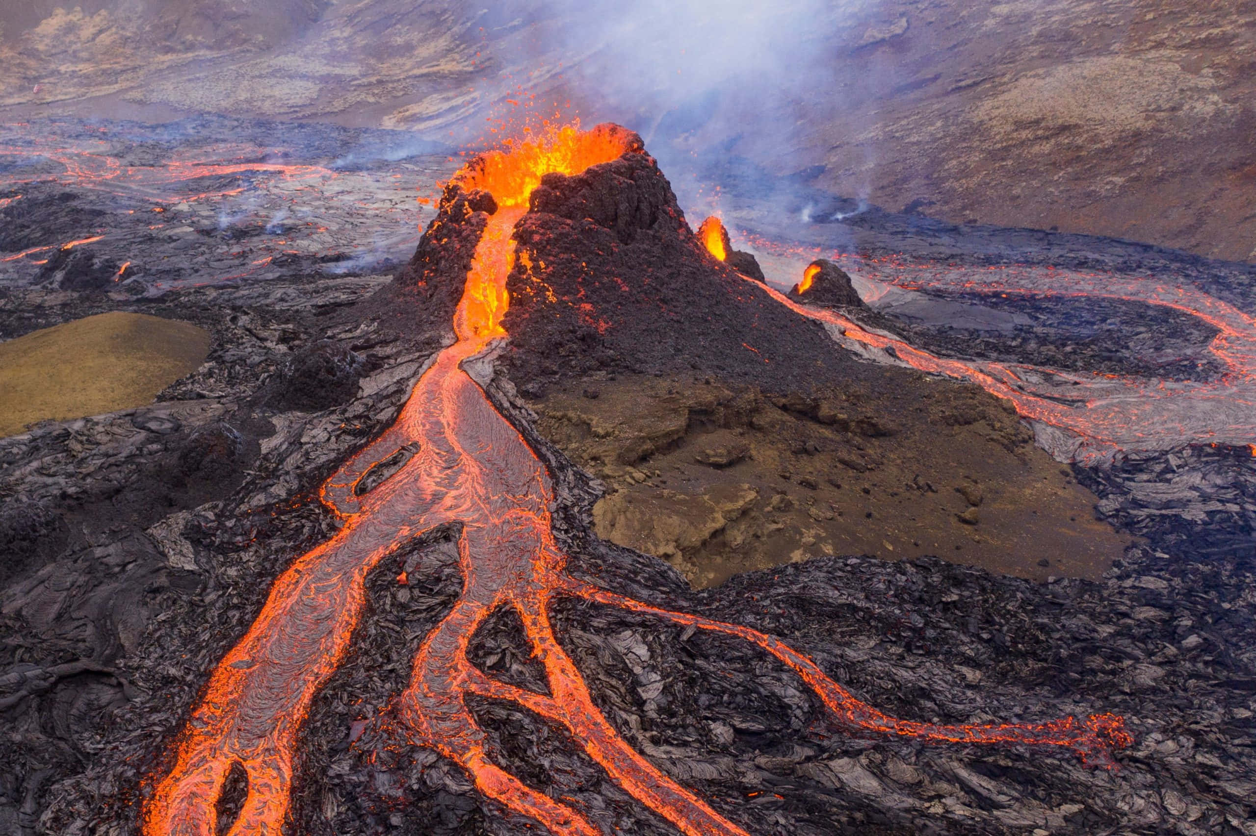 Volcano 桌布 壁紙 — 火山爆發在昏暗的天空下，將熾熱的橙色熔岩噴灑在崎嶇的黑色岩層上。