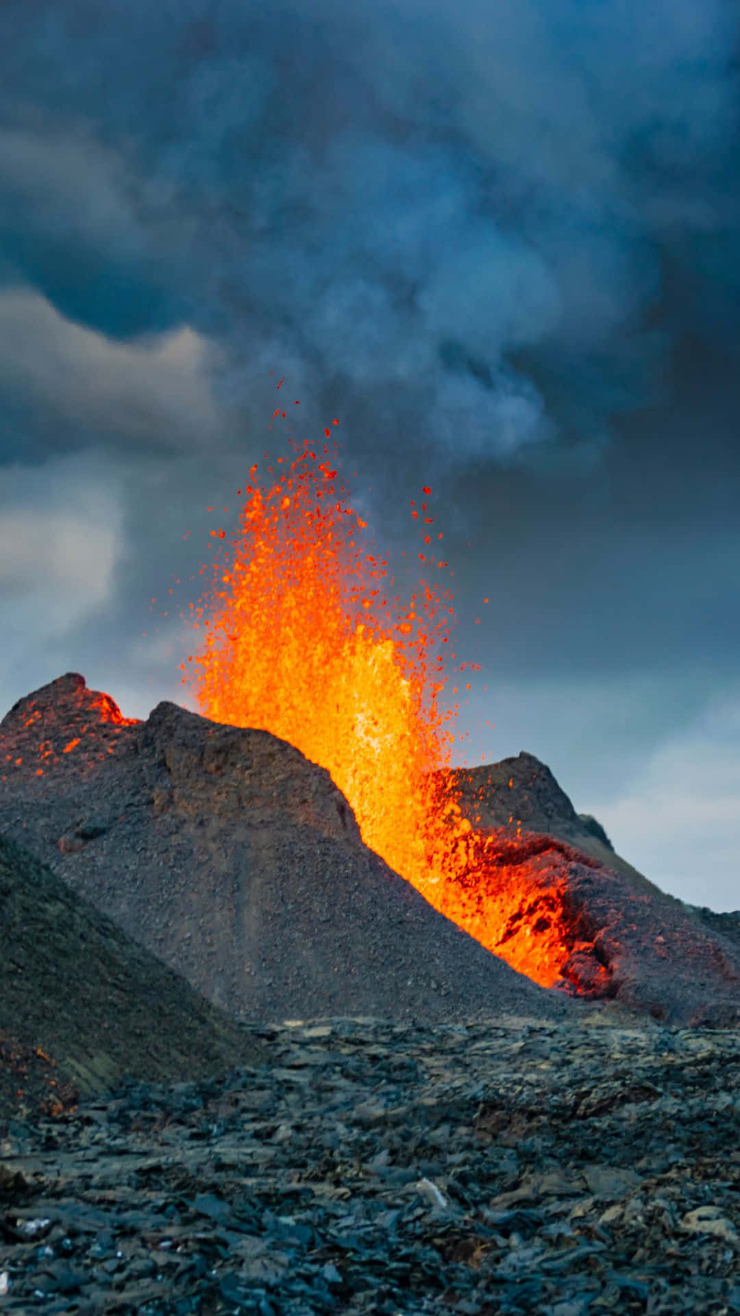 冰島 桌布 壁紙 — 火山爆發將熾熱的橙色熔岩和火山灰噴向風暴般的黑暗天空，下方是崎嶇的黑色岩石地形。