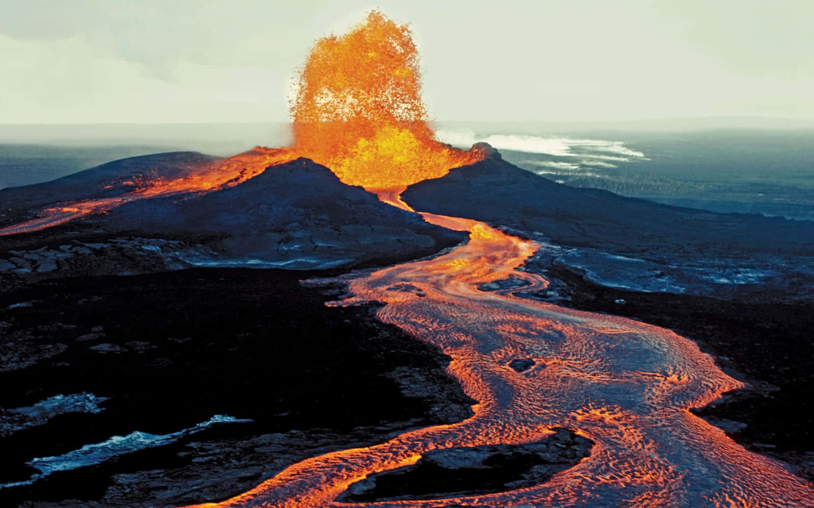 Volcano 桌布 壁紙 — 火山猛烈爆發，噴出熾熱的橙色熔岩，像液體火焰一樣流過黑暗的火山景觀。