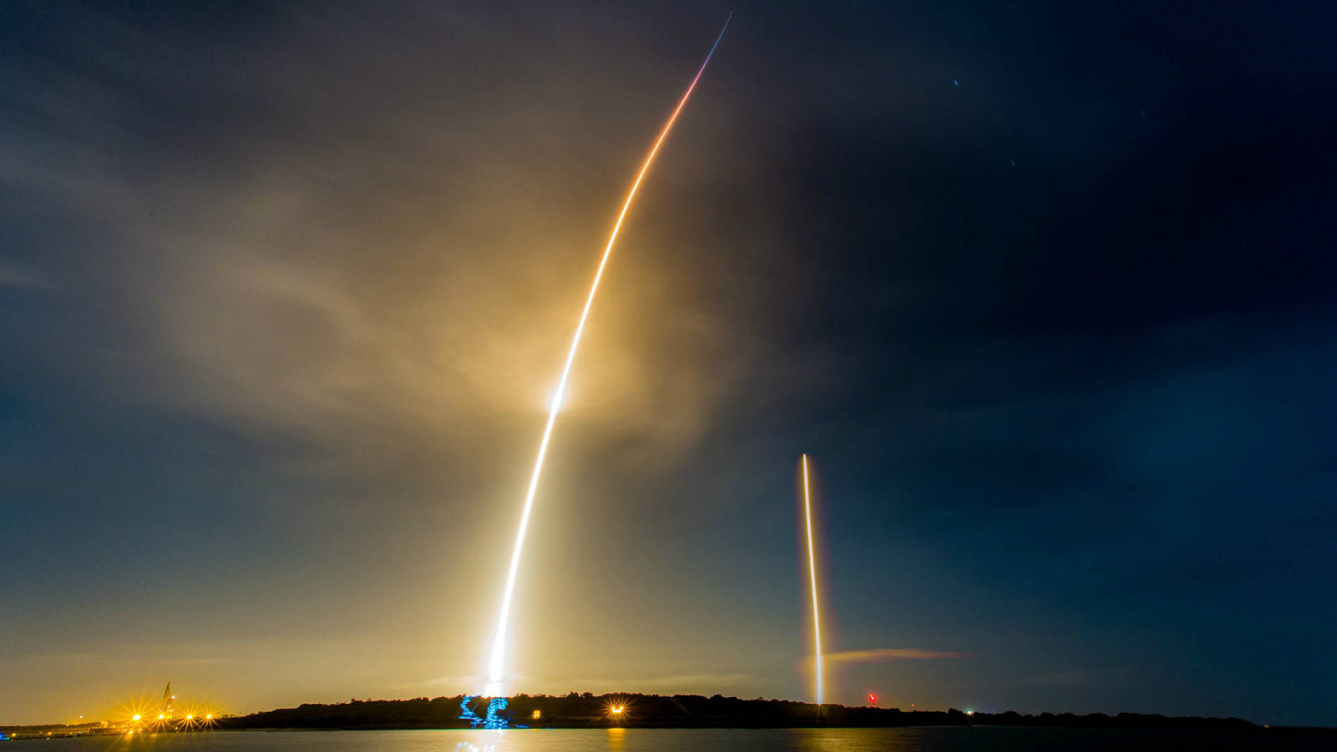 SpaceX 桌布 壁紙 — 火箭在夜間升空，在黑暗的天空中留下燦爛的金色軌跡，下方有薄雲和遠處的燈光。