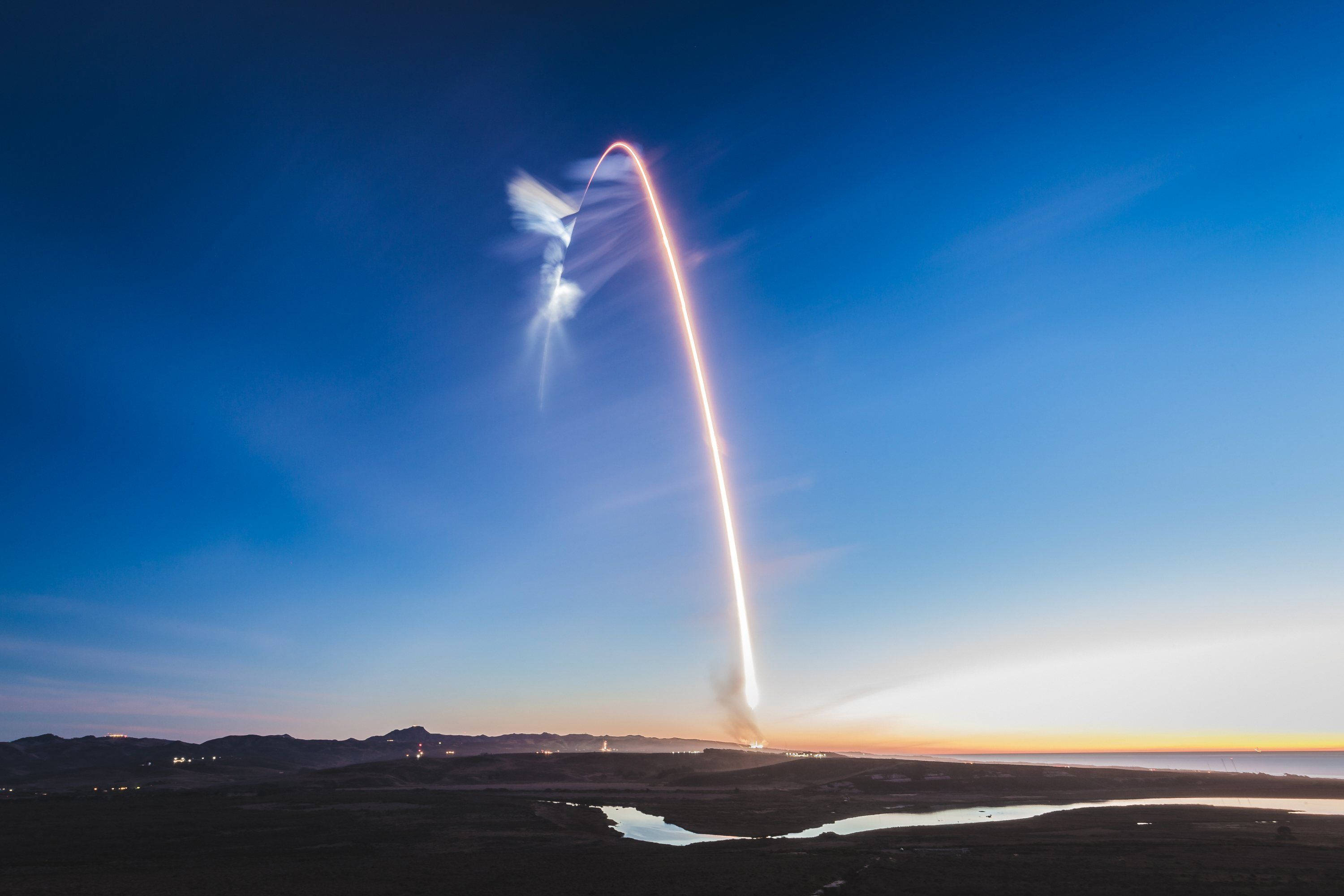 SpaceX 桌布 壁紙 — 火箭在暮色天空中劃出燦爛的弧線上升，越過沙漠景觀，遠處有山脈和蜿蜒的河流。