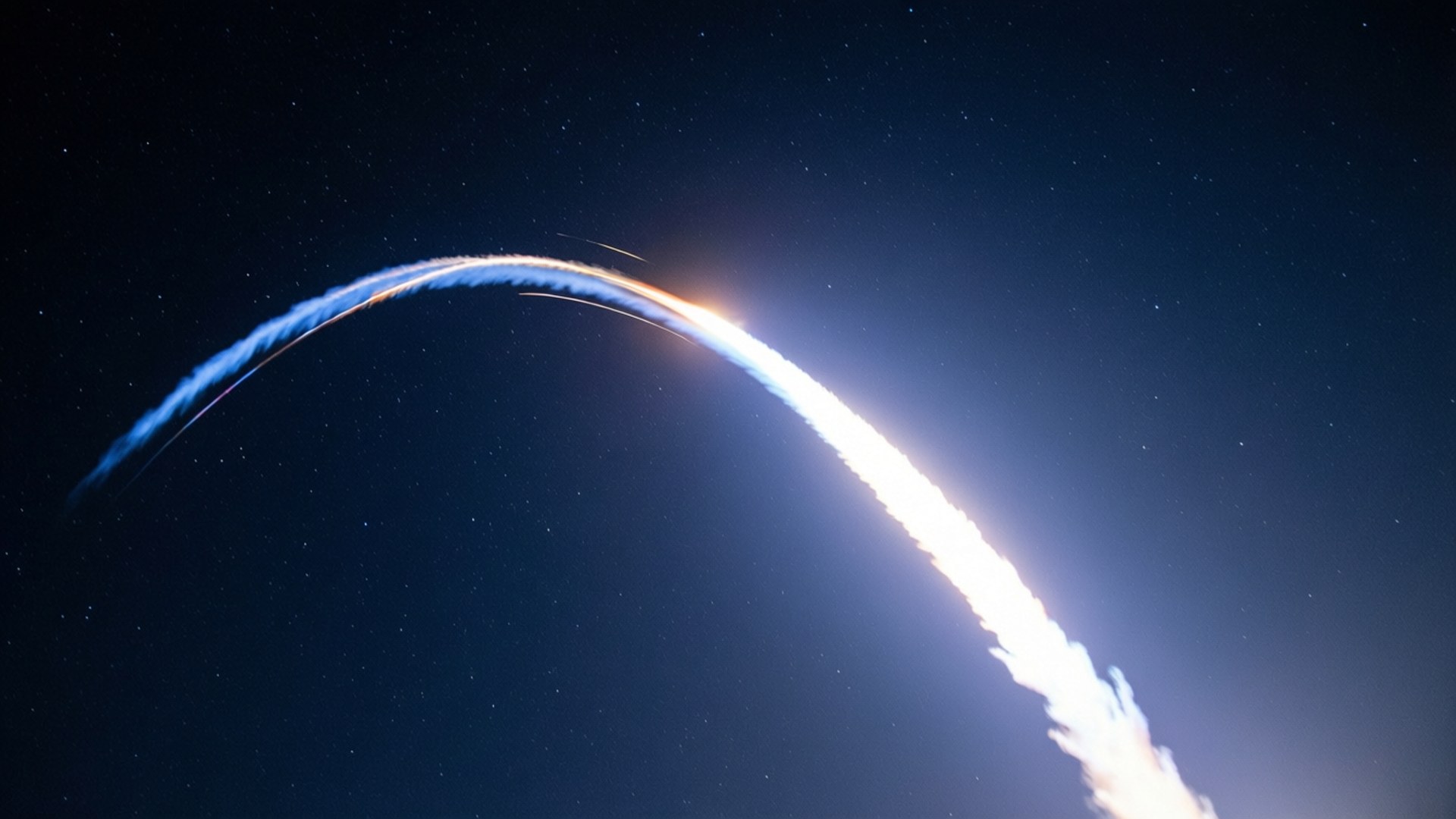 火箭軌跡縮時攝影 壁紙 — 火箭在繁星點點的夜空中以燦爛的弧線上升，留下發光的藍白色軌跡。