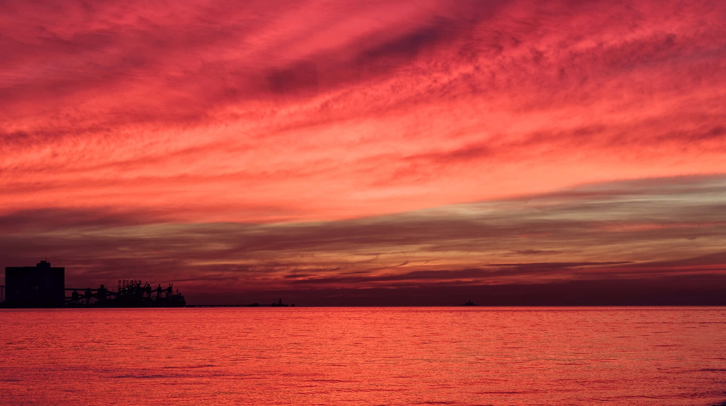海灘日落 桌布 壁紙 — 火紅的夕陽染紅了天空和海洋，地平線上矗立著海岸建築的黑色剪影。