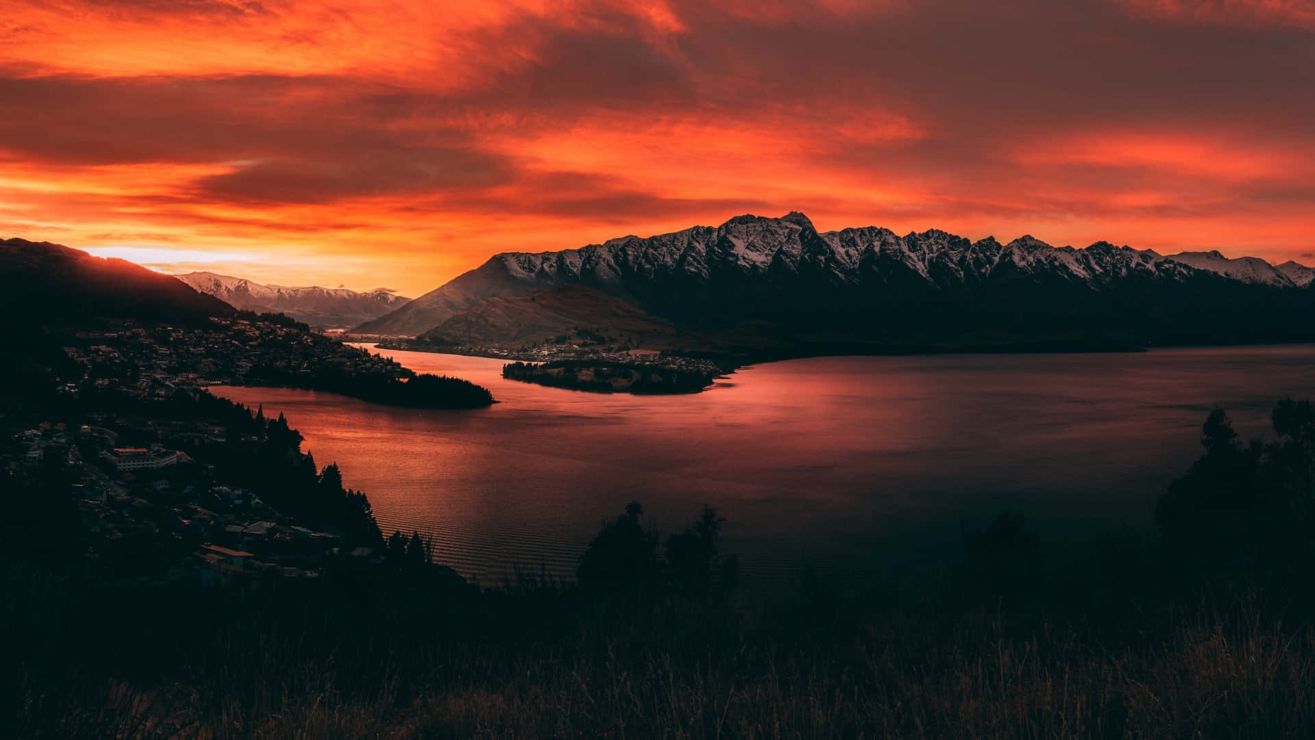 美學 桌布 壁紙 — 火紅的橙色夕陽染紅了天空，倒映在平靜的湖面上，四周是白雪覆蓋的山脈和一個小鎮。