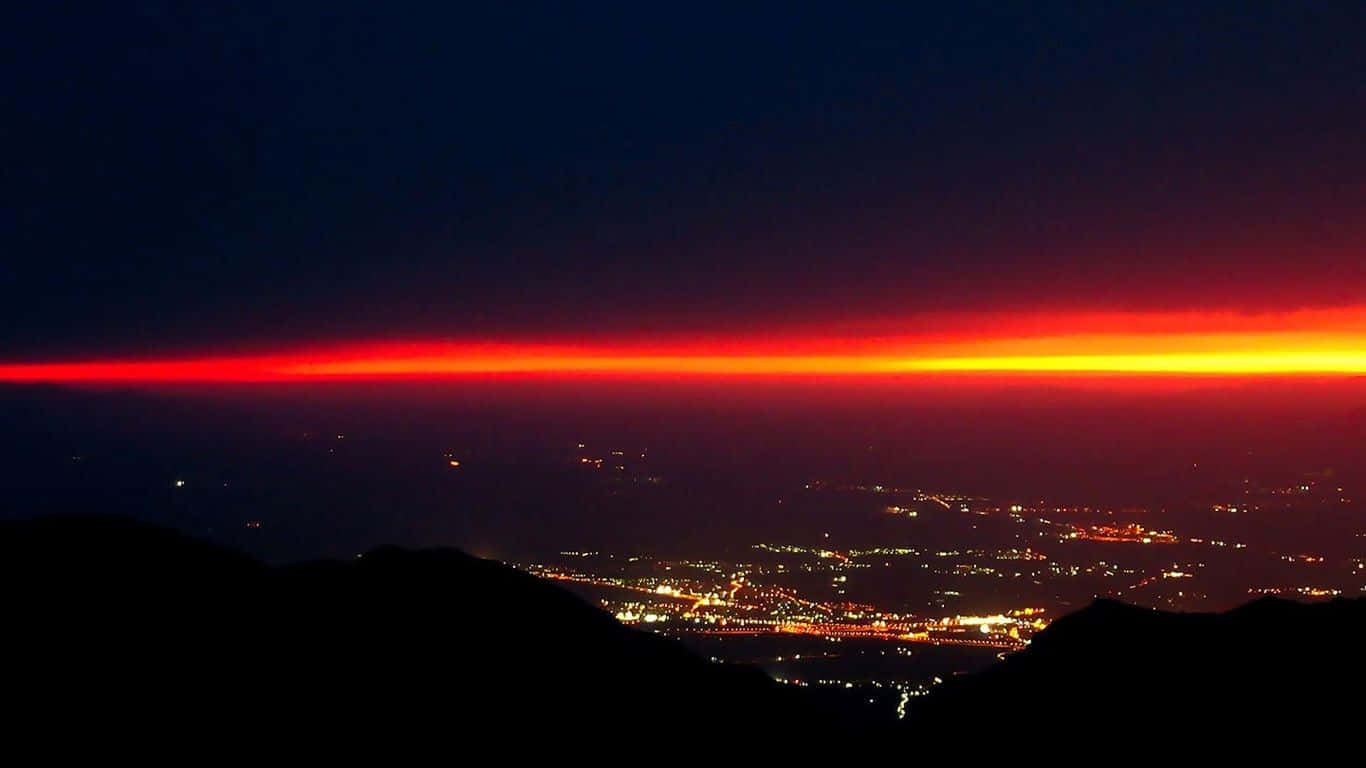 筆電 桌布 壁紙 — 火紅與橙色的地平線在夜間廣闊的城市景觀上方發光，前景有黑暗的輪廓山丘。