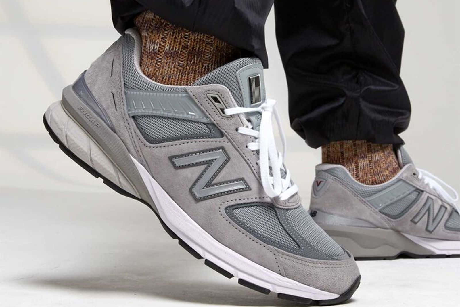 New Balance 桌布 壁紙 — 灰色New Balance運動鞋特寫，搭配白色鞋帶，穿著圖案襪子和黑色褲子，在淺色地板上。