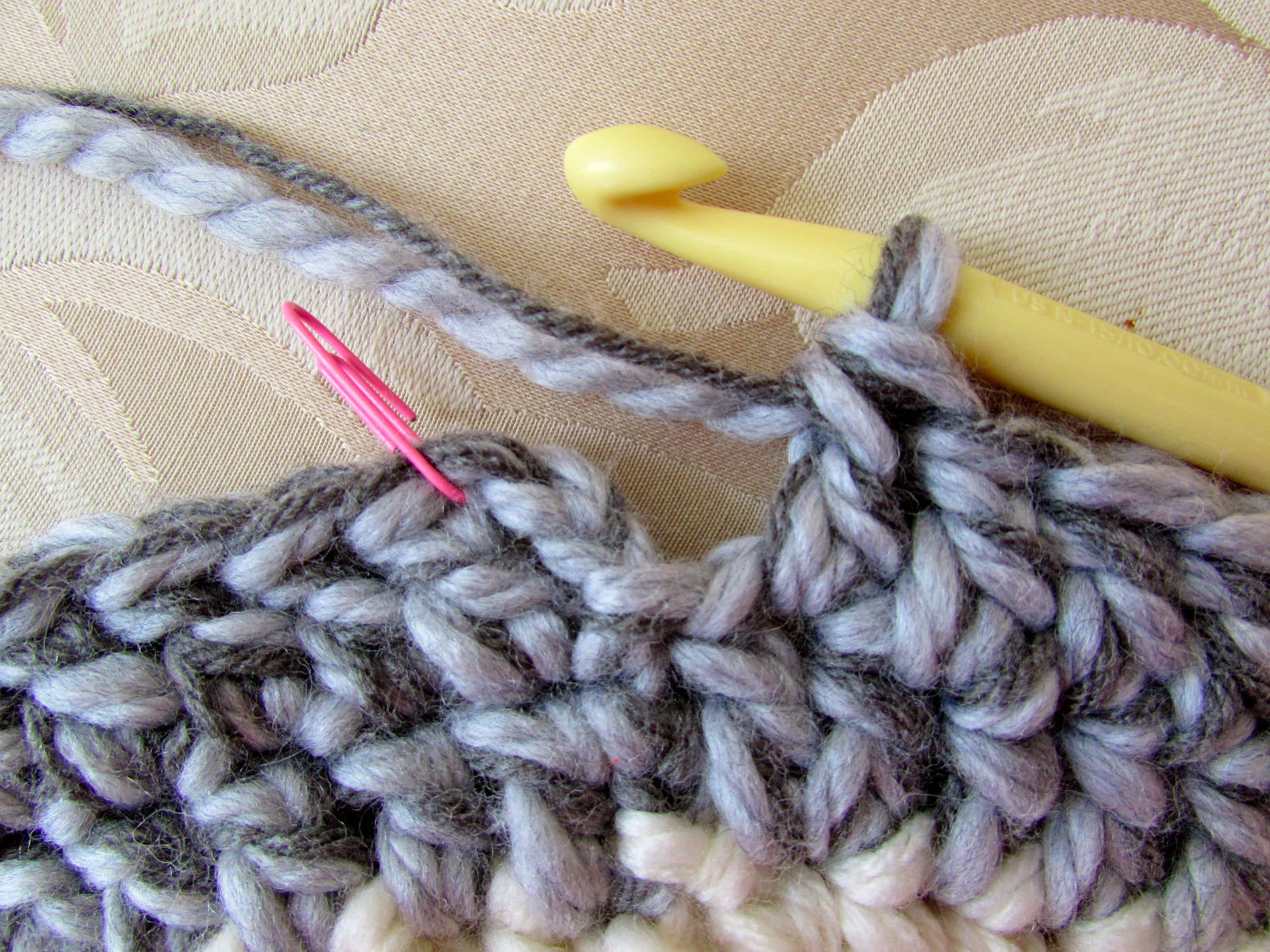Crochet 桌布 壁紙 — 灰色和奶油色鉤針編織布料的特寫，黃色鉤針和粉紅色針跡標記放在有紋理的米色布料上。