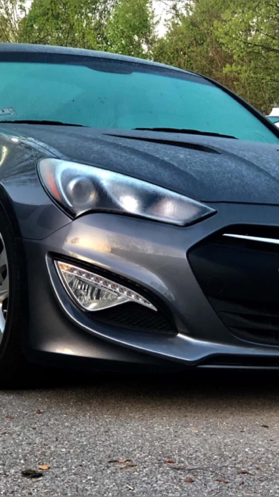 Hyundai Genesis 桌布 壁紙 — 灰色時尚跑車前端的特寫，配有銳利頭燈和光澤引擎蓋，停在碎石上，附近有綠樹。