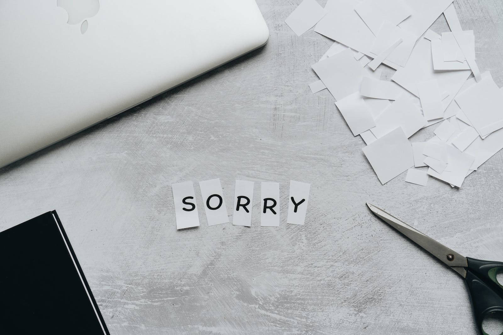 Sorry 桌布 壁紙 — 灰色桌子上，剪刀和筆記型電腦旁邊，用剪下的紙片拼出「sorry」這個詞。