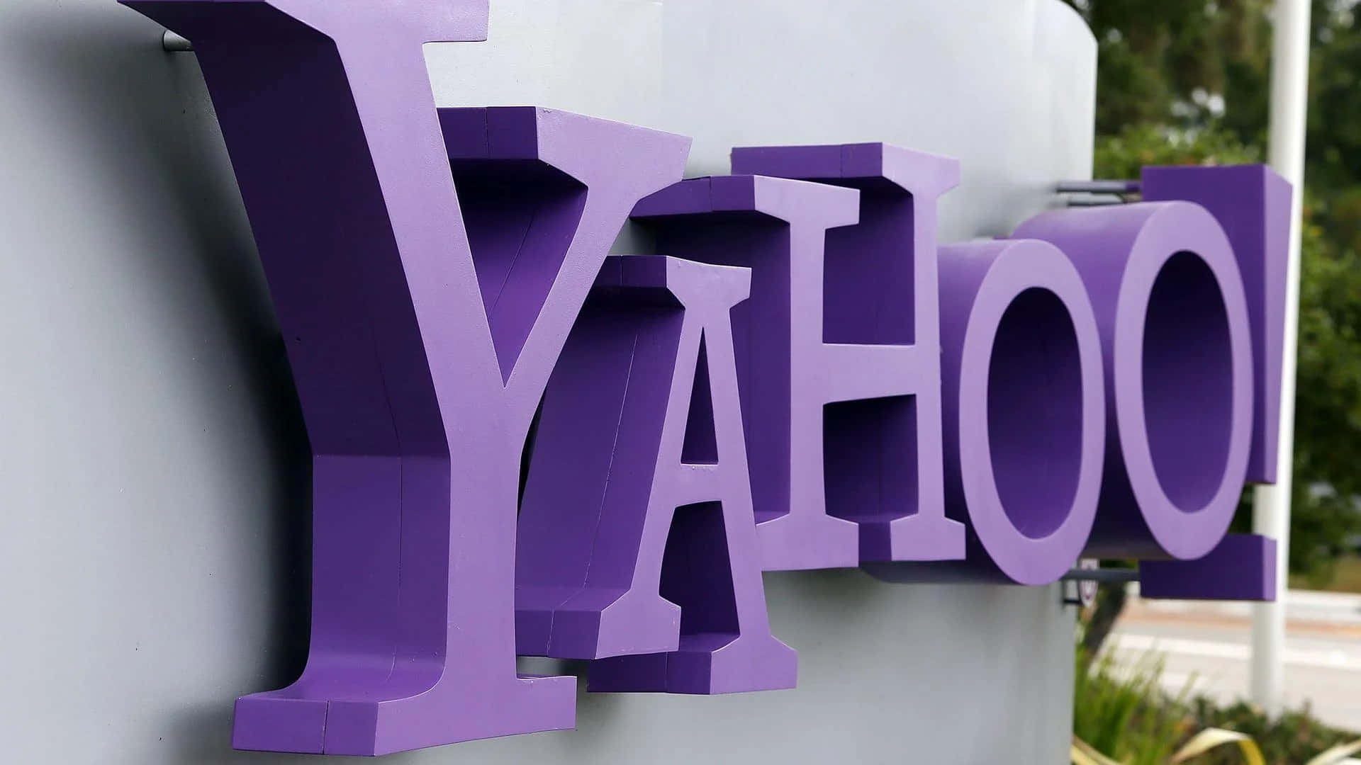 Yahoo 桌布 壁紙 — 灰色牆上安裝的大型紫色 Yahoo 標誌特寫，背景有綠色植物。