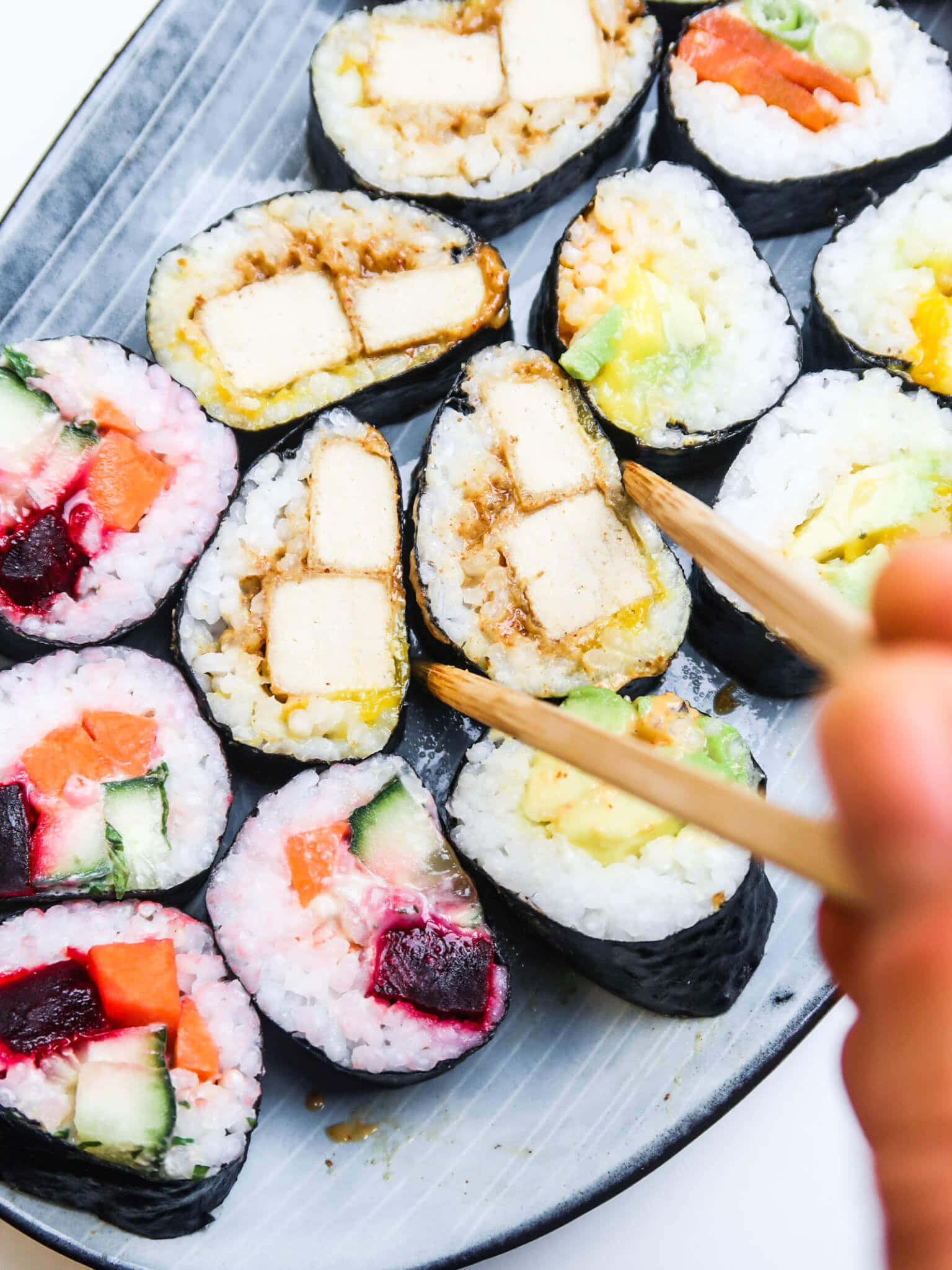 Sushi 桌布 壁紙 — 灰色盤子上色彩繽紛的壽司捲特寫，筷子正夾起一個。