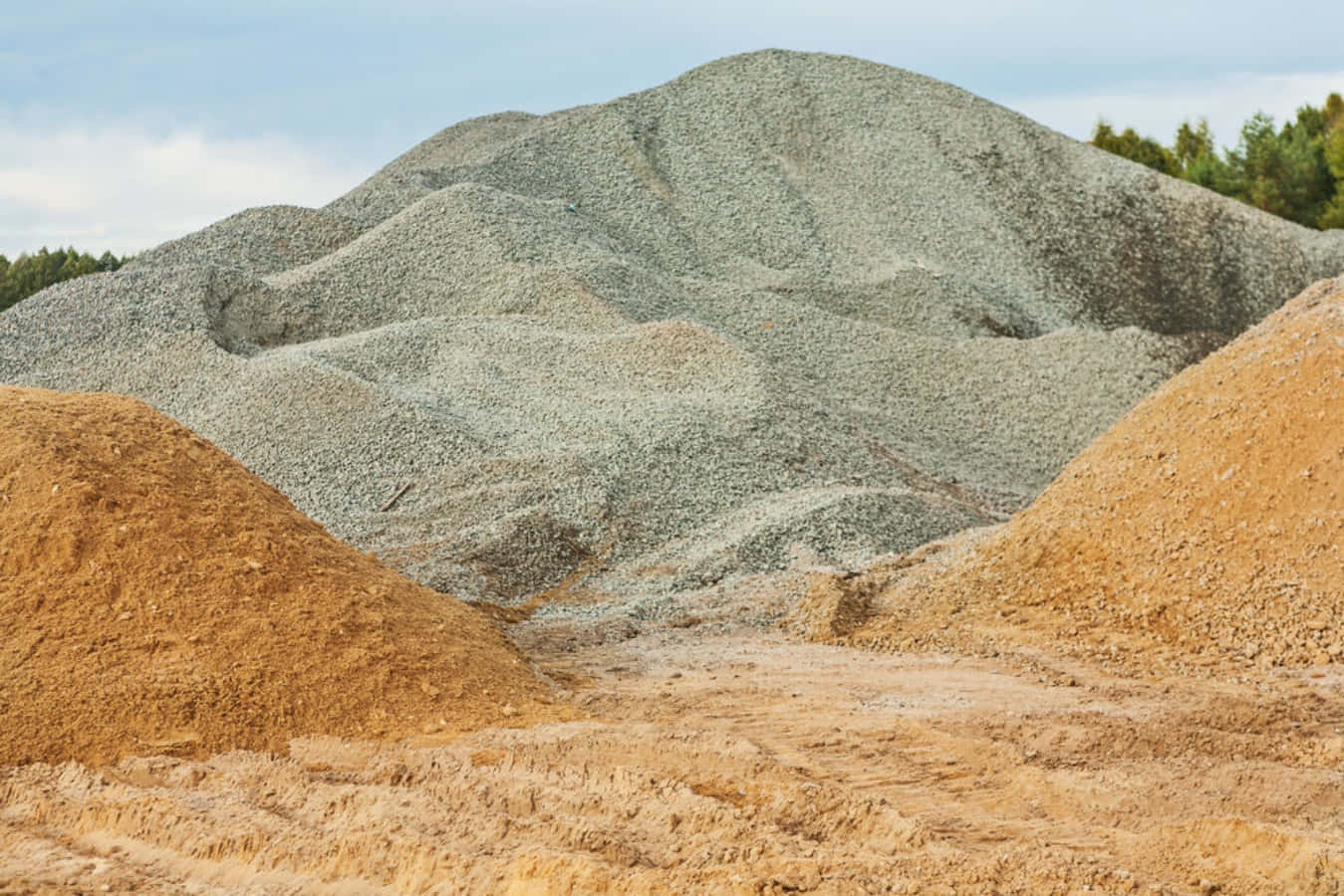 sand 桌布 壁紙 — 灰色碎石和橙色沙子的巨大土堆主導著一個陽光明媚的建築工地，背景有樹木。