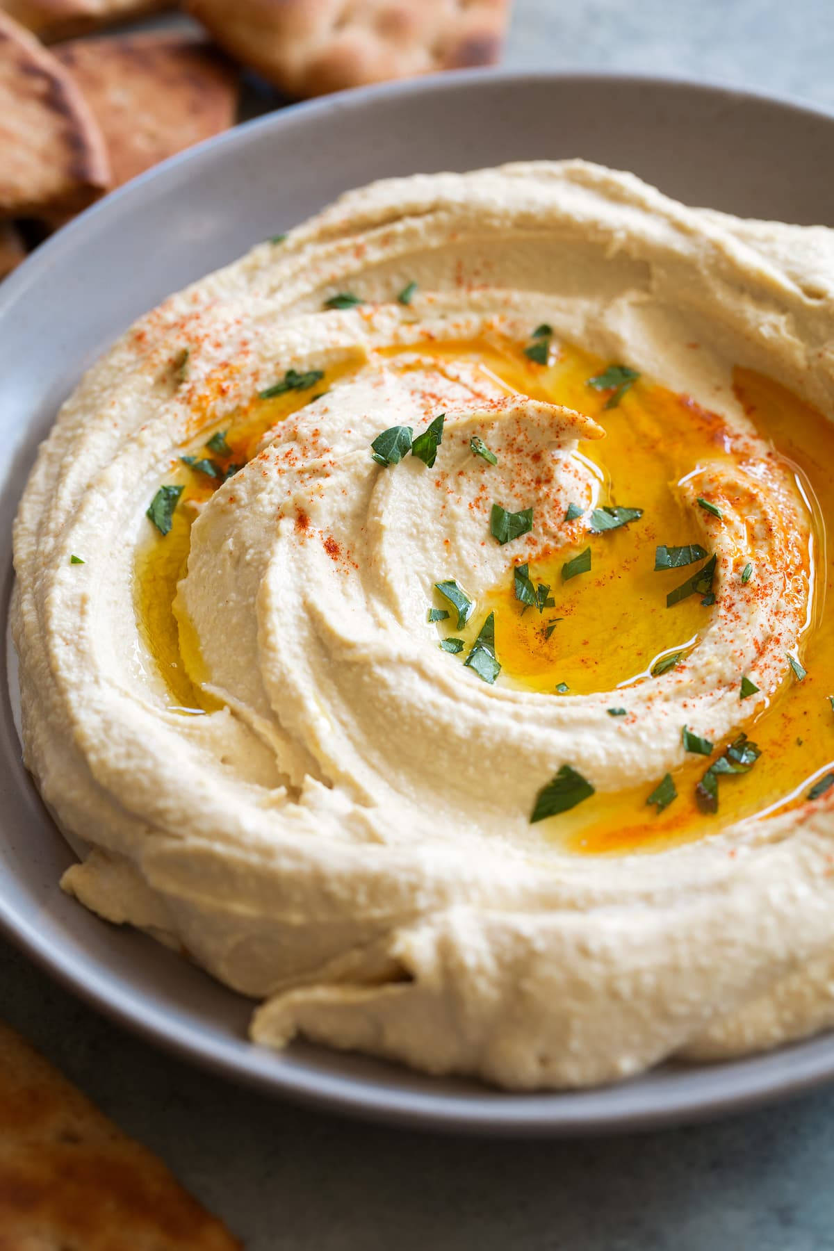 Hummus 桌布 壁紙 — 灰色碗中的奶油鷹嘴豆泥漩渦，上面點綴著金黃色橄欖油、紅椒粉和新鮮歐芹。