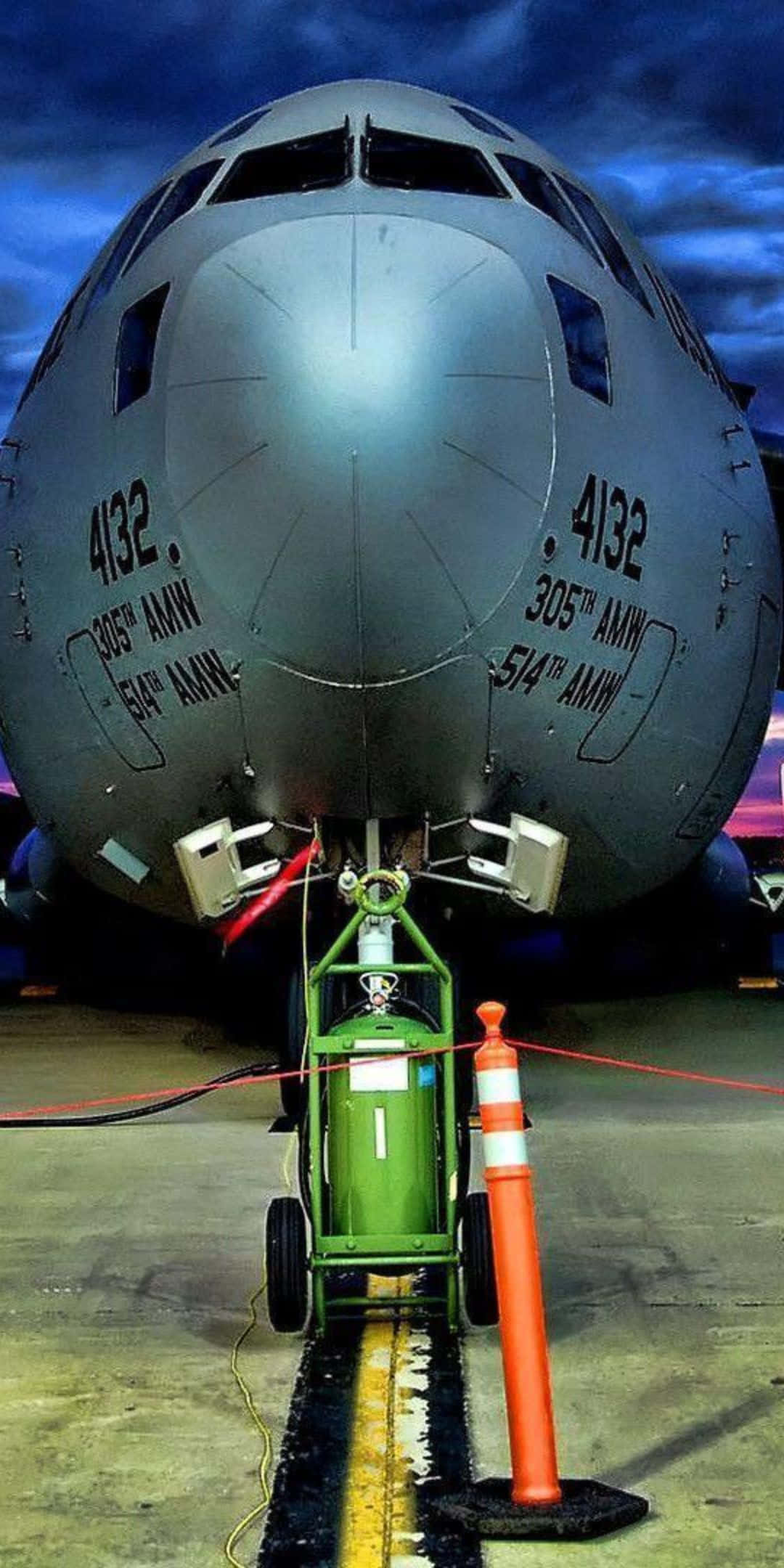 Pixel 3 Plane 桌布 壁紙 — 灰色軍用運輸機機頭近距離特寫，帶有維修設備和橙色錐體，在戲劇性的暮色天空下的停機坪上。