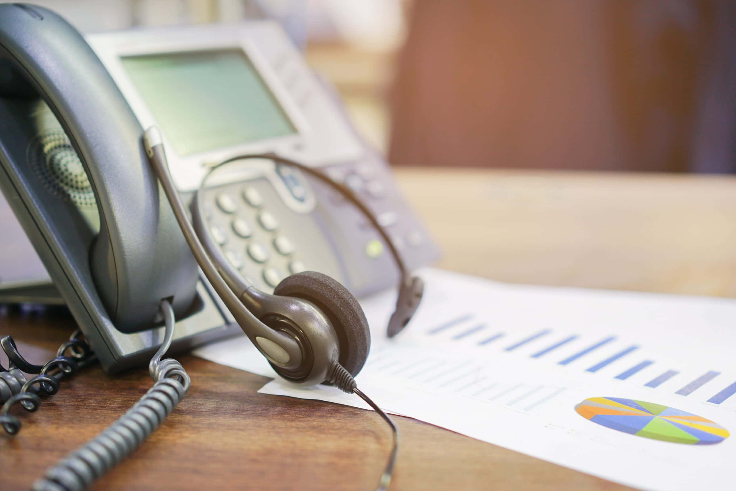 Telemarketing 桌布 壁紙 — 灰色辦公室電話和耳機放在木桌上，旁邊是色彩繽紛的商業圖表的特寫。