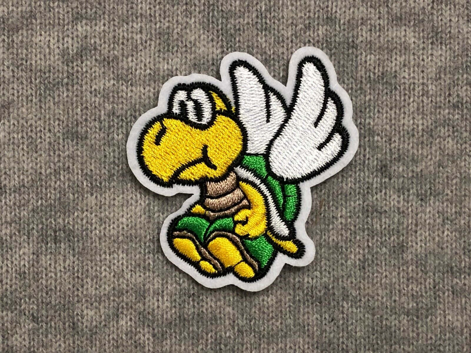 Koopa Troopa 桌布 壁紙 — 灰色針織布料上，一個色彩繽紛的刺繡補丁，圖案為一隻黃色飛天龜角色，帶有白色翅膀。