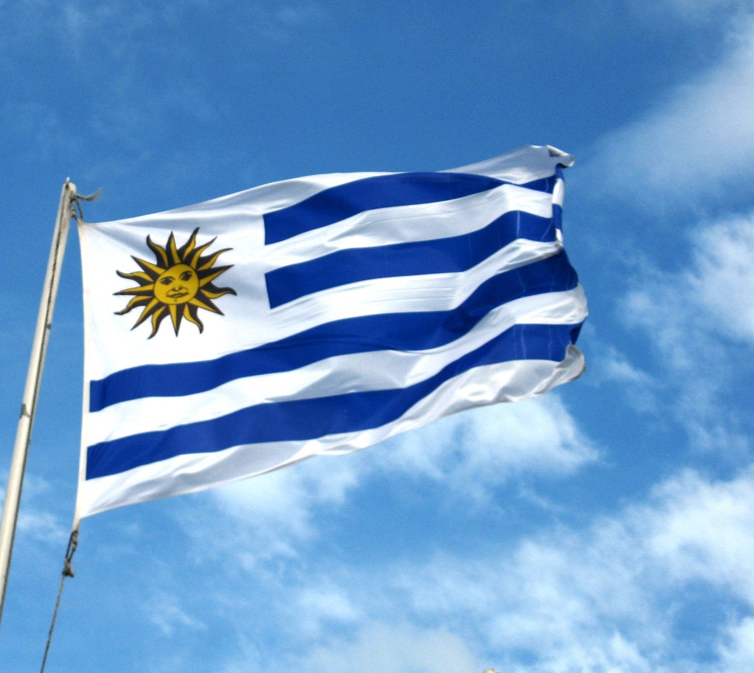 Uruguay 桌布 壁紙 — 烏拉圭國旗，帶有藍白條紋和金色太陽標誌，在藍天白雲中自豪地飄揚。