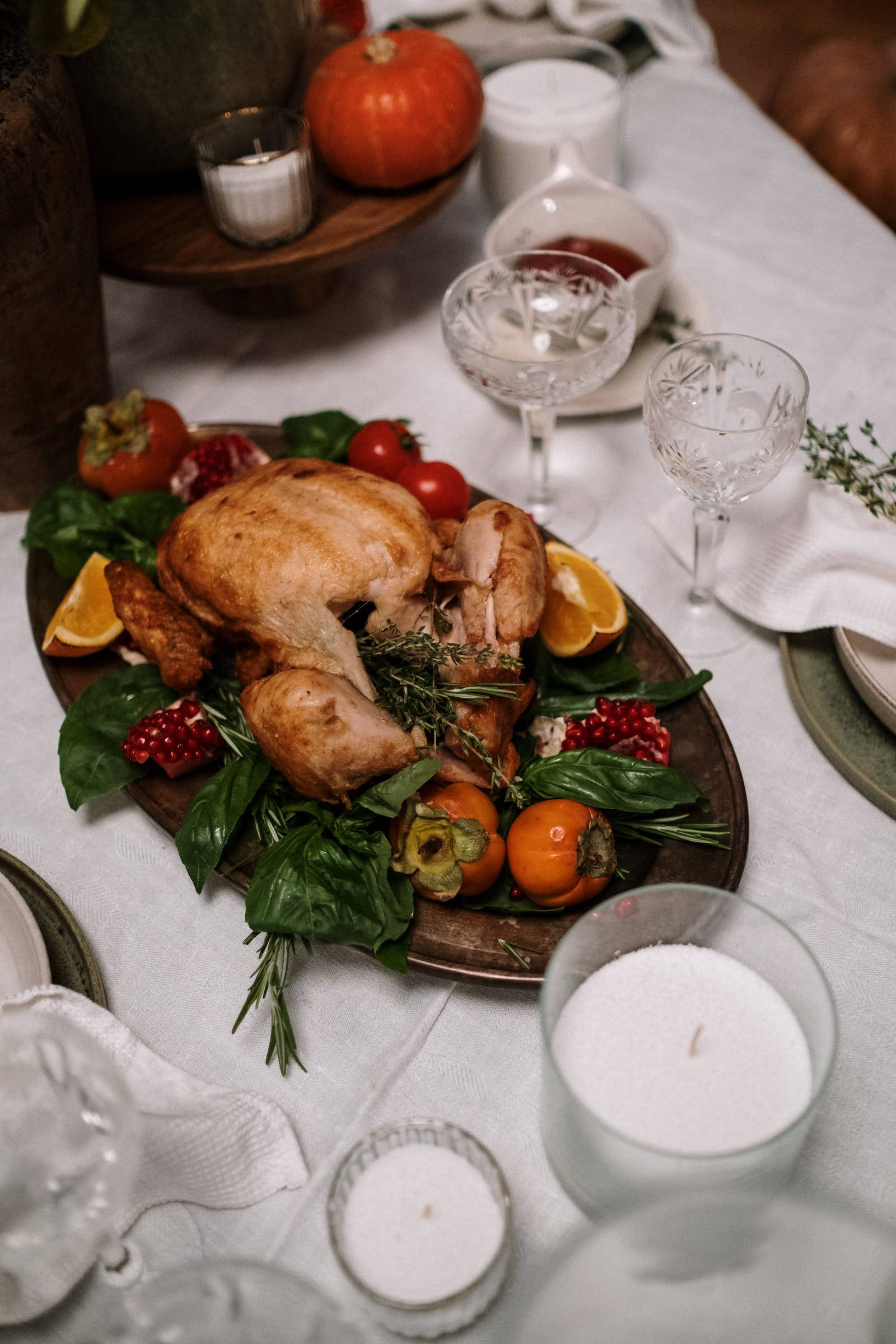 Thanksgiving Day 桌布 壁紙 — 烤火雞作為中心擺放在木盤上，周圍有柿子、石榴和香草，放在白色桌布上，配有蠟燭和玻璃杯。
