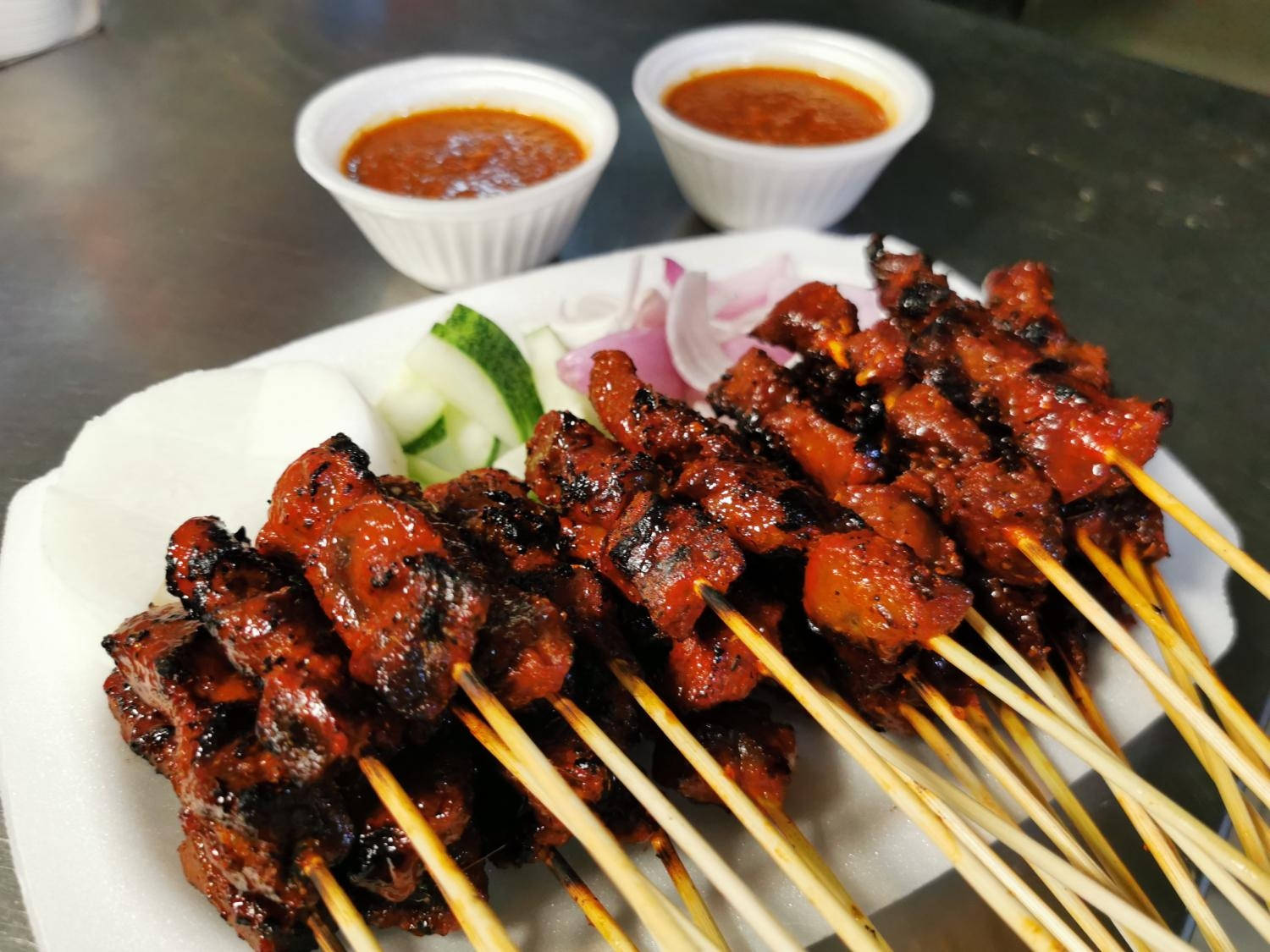 Satay 桌布 壁紙 — 烤肉串閃耀著焦糖化的光澤，旁邊是新鮮的小黃瓜和洋蔥片，還有兩小碗鮮紅色的醬汁。