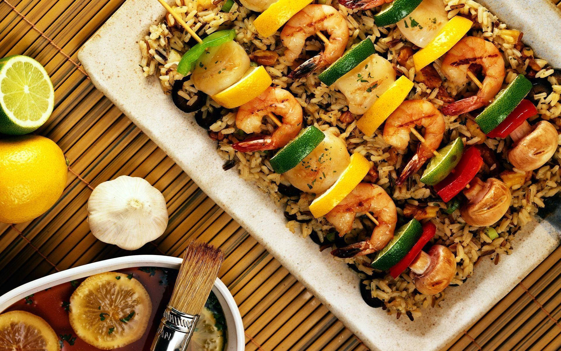 Delicious Food 桌布 壁紙 — 烤蝦和蔬菜串放在野生米飯上，置於竹蓆上，旁邊有新鮮檸檬和大蒜。