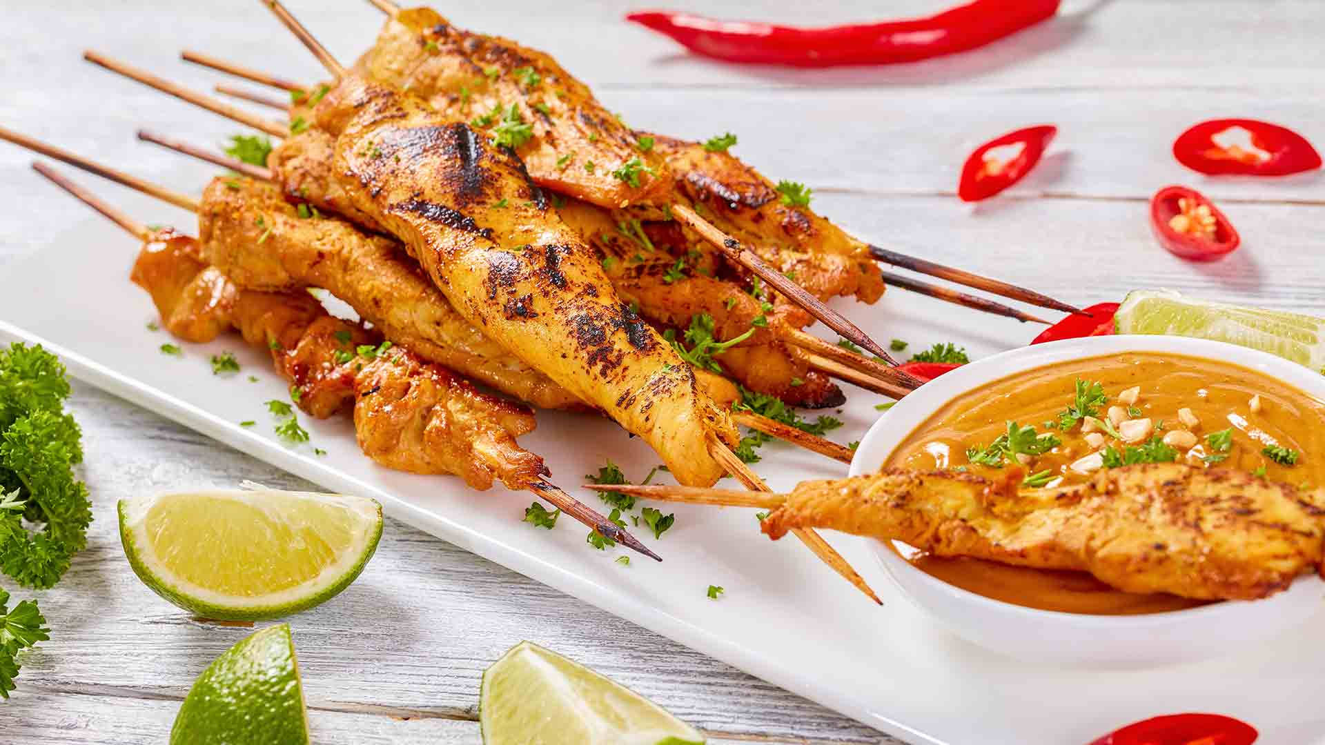 Satay 桌布 壁紙 — 烤雞肉沙爹串配花生醬，飾以香菜、萊姆角及紅辣椒，置於白色木桌上。