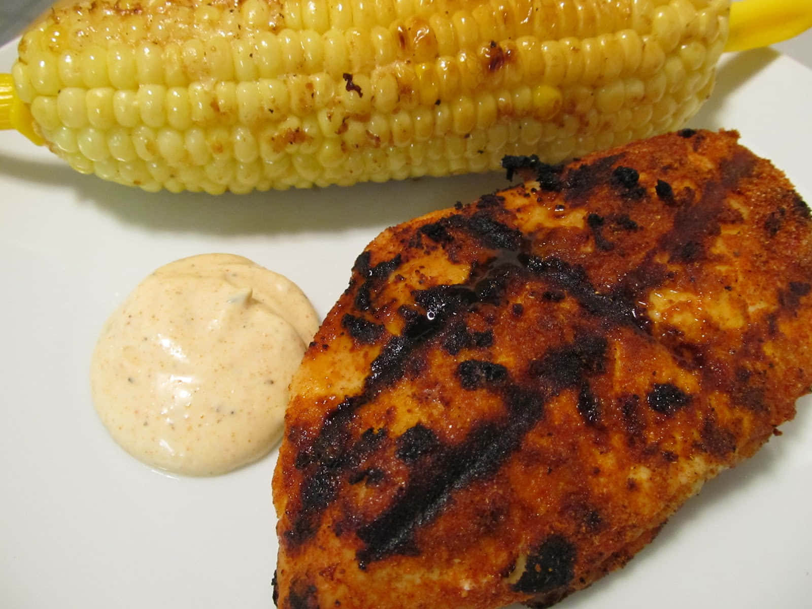 Blackened Chicken 桌布 壁紙 — 烤雞胸肉帶有烤痕，旁邊放著一條玉米和奶油醬汁，盛在白色盤子上。