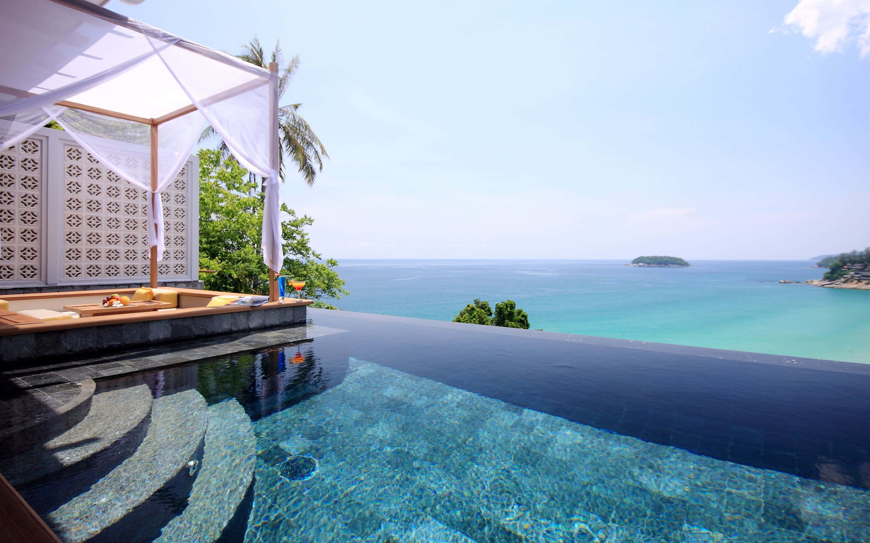 Thailand Beach 桌布 壁紙 — 無邊際泳池俯瞰著碧綠色的海洋，設有白色遮陽篷休息區和棕櫚樹，在明亮的藍天下。