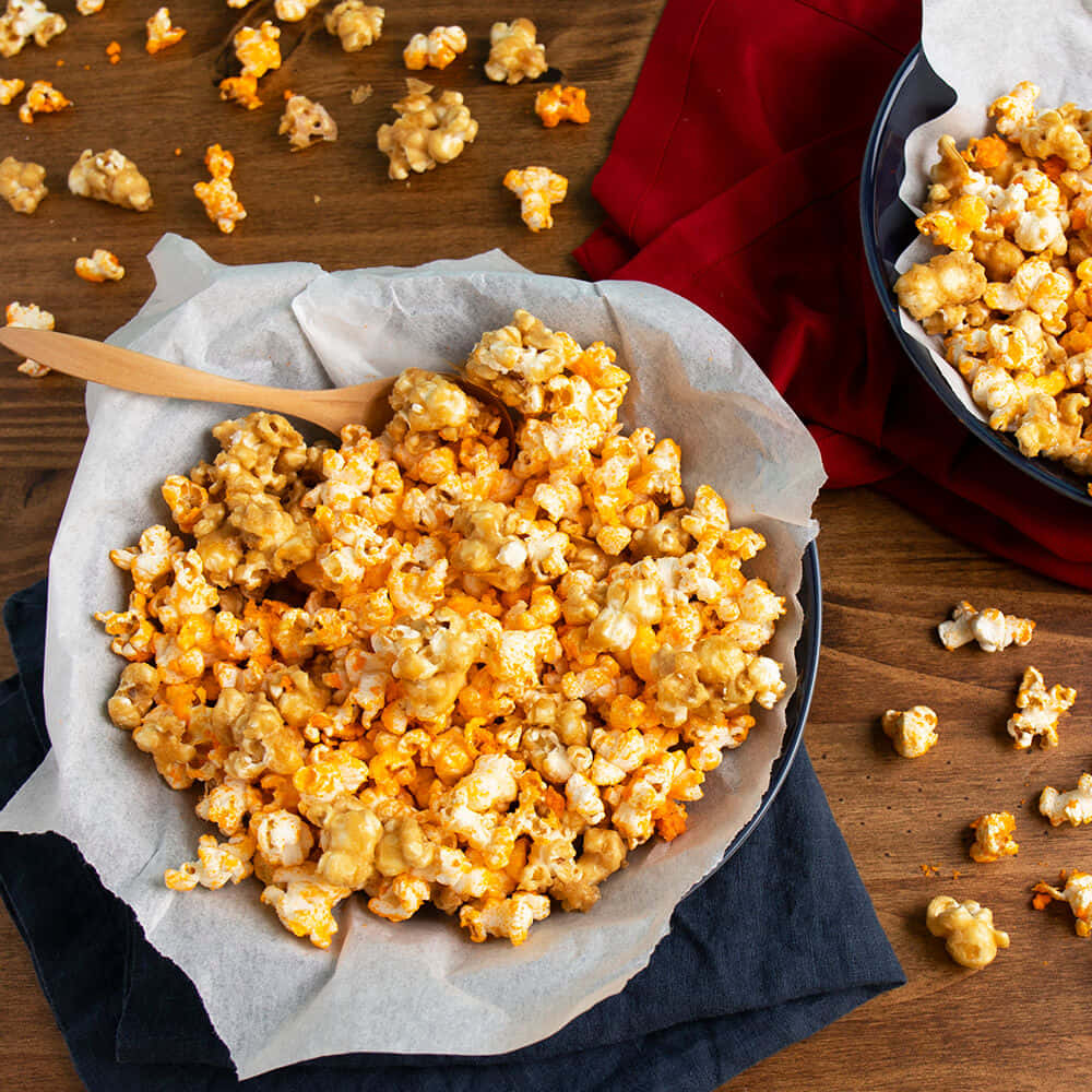 Popcorn 桌布 壁紙 — 焦糖爆米花從鋪有烘焙紙的深色碗中溢出，落在鋪有紅色和海軍藍餐巾的木桌上。