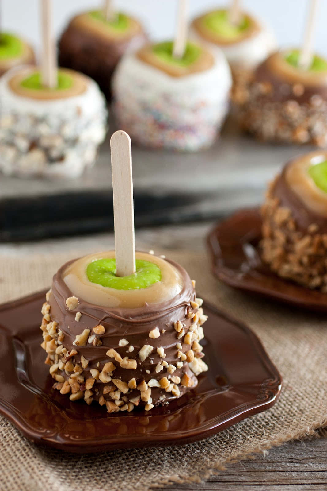 Caramel Apples 桌布 壁紙 — 焦糖蘋果特寫，外層裹上巧克力和碎堅果，放在深色盤子上，背景為麻布。
