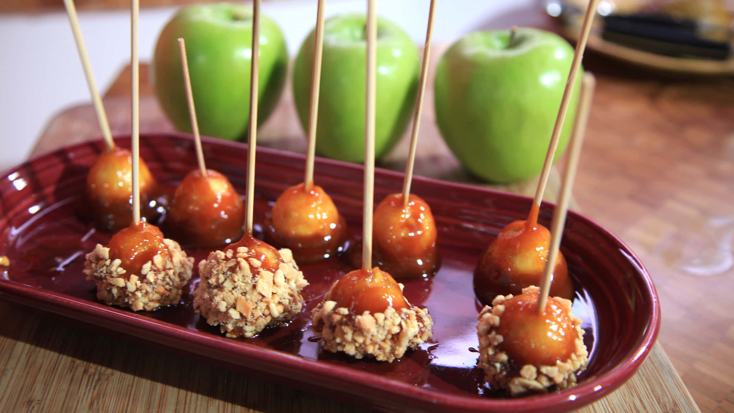 Caramel Apples 桌布 壁紙 — 焦糖蘋果裹上碎堅果，插在棒上，排列在光亮的紅色托盤上，背景有完整的青蘋果。