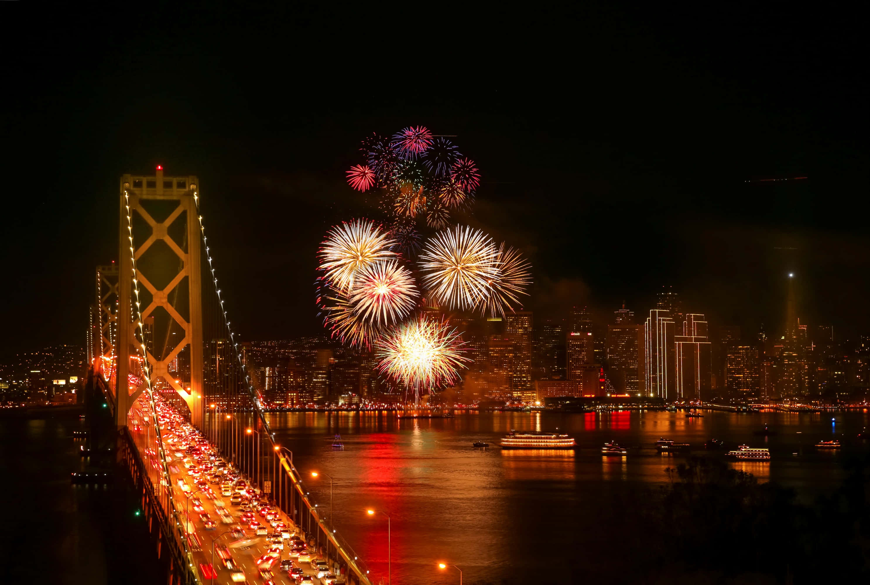 New Years Eve 桌布 壁紙 — 煙火在一座亮燈的橋樑和城市天際線上爆炸，下方交通在漆黑的夜晚中流動。