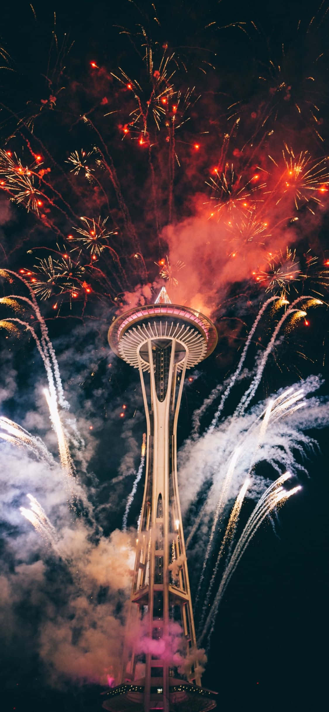 iPhone X Seattle 桌布 壁紙 — 煙火在夜晚圍繞著被照亮的太空針塔爆炸，呈現鮮豔的紅色和白色。