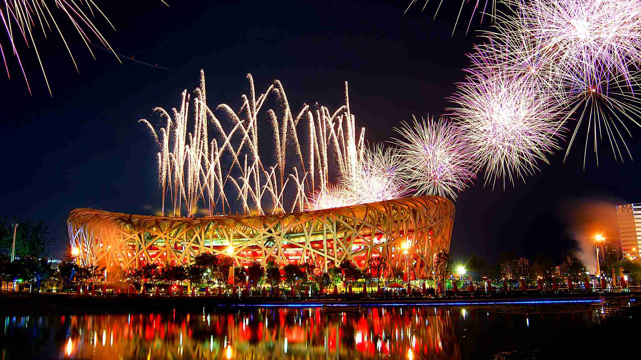Beijing 桌布 壁紙 — 煙火在夜晚點亮的鳥巢體育場上空爆炸，下方水面有鮮豔倒影。