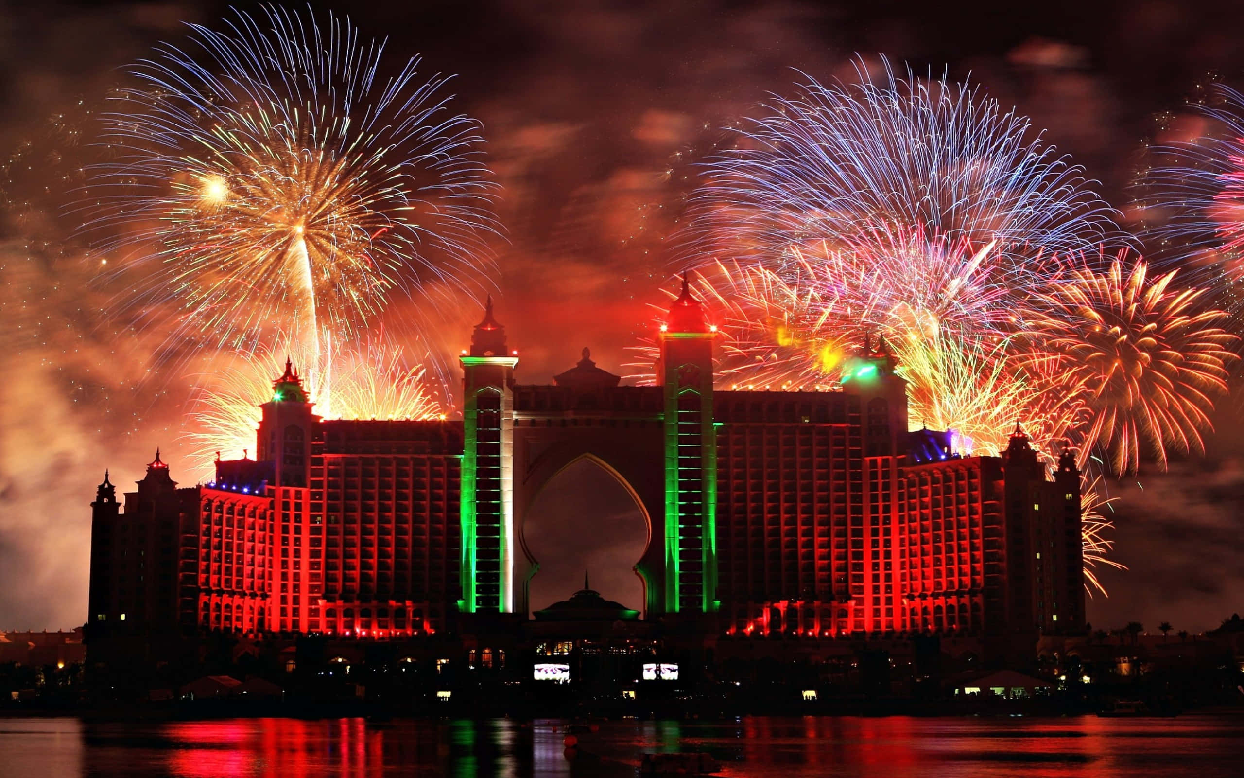 Fireworks 桌布 壁紙 — 煙火在夜間於一座宏偉的燈光酒店上空綻放，色彩繽紛，倒映在水面上。