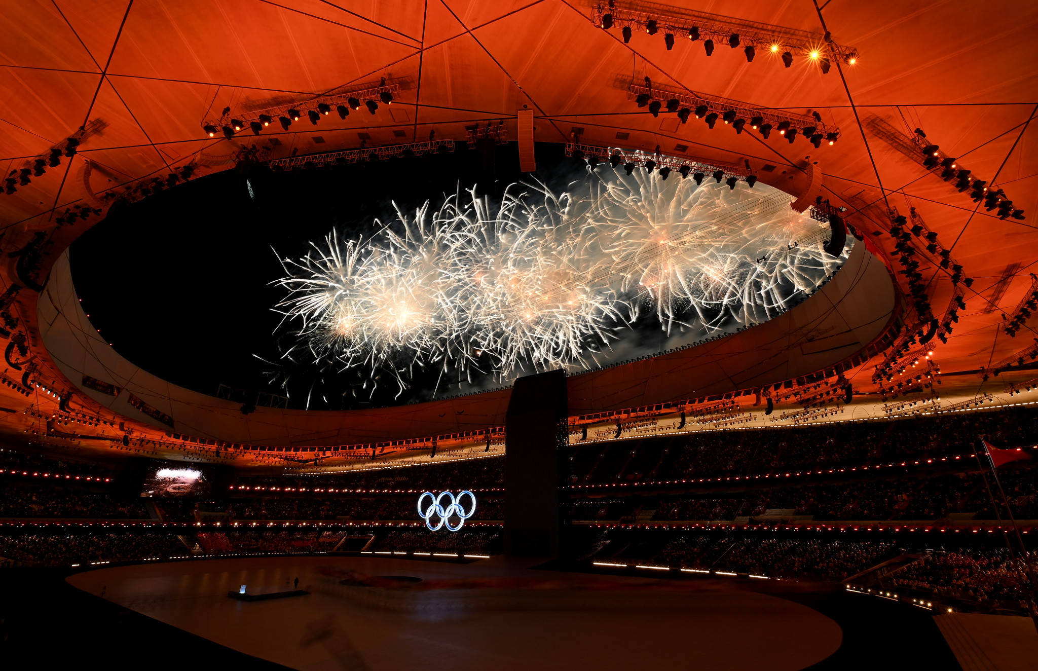 Winter Olympics 桌布 壁紙 — 煙火在擠滿人的奧林匹克體育場上空爆炸，下方是發光的橙色屋頂和奧林匹克五環。