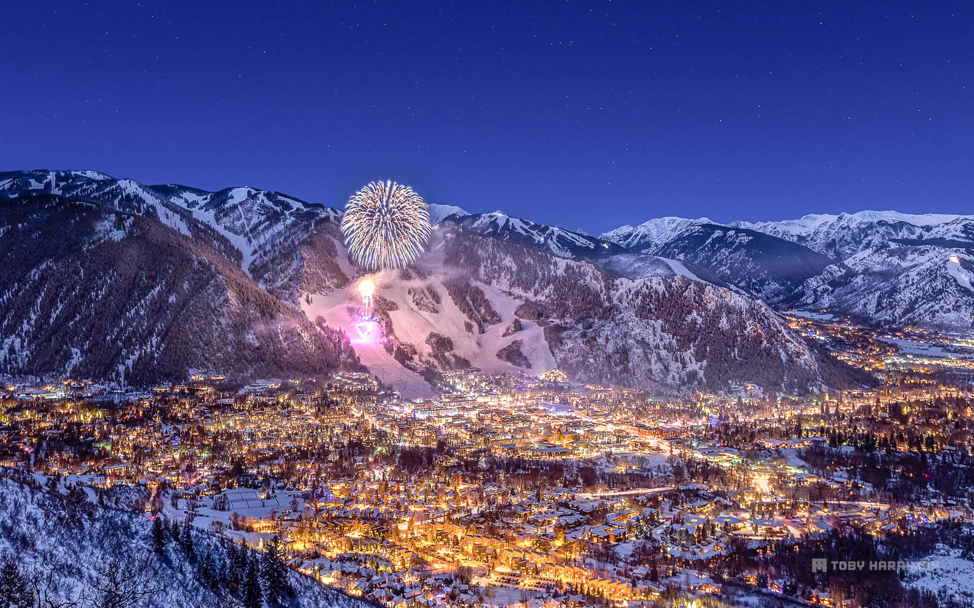 Aspen Colorado 桌布 壁紙 — 煙火在星空下白雪覆蓋的山城上空綻放，溫暖的燈光閃爍。